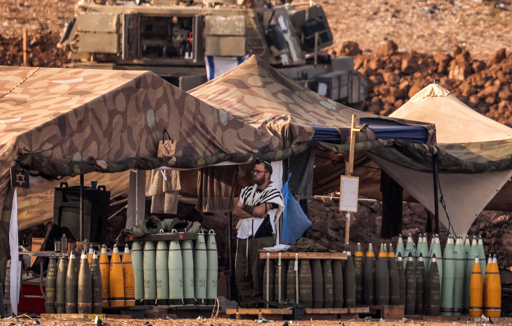 این تصویر نمایی از یک منطقه نظامی را نشان میدهد. در اینجا، چادرهای نظامی با طرح camouflage قرار دارند و به نظر میرسد که وسایل نظامی و مهمات در اطراف آنها وجود داشته باشد. فردی با لباس خاص، در کنار چادرها ایستاده است. همچنین، تعدادی مهمات در رنگهای مختلف به وضوح قابل مشاهده است. در پسزمینه، تعدادی وسیله نقلیه نظامی نیز به چشم میخورد. این صحنه حس فعالیت نظامی و آمادهسازی را منتقل میکند.