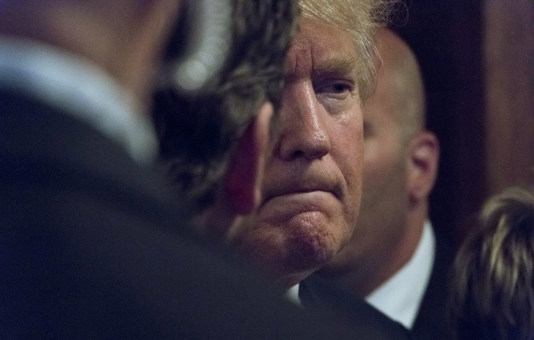 در این تصویر، یک مرد با موهای روشن و کمی حالت خمیده سر دیده میشود. او در حالتی contemplative به نظر میرسد و گونهها و خطوط صورتش مشخص است. در اطراف او چندین فرد دیگر مشاهده میشوند که به نظر میرسد در حال گفتگو یا مراقبت از او هستند. پسزمینه تصویر تاریک و مبهم است، که تمرکز را بر روی چهره او بیشتر میکند. این تصویر احساس جدیت و تفکر عمیق را منتقل میکند.