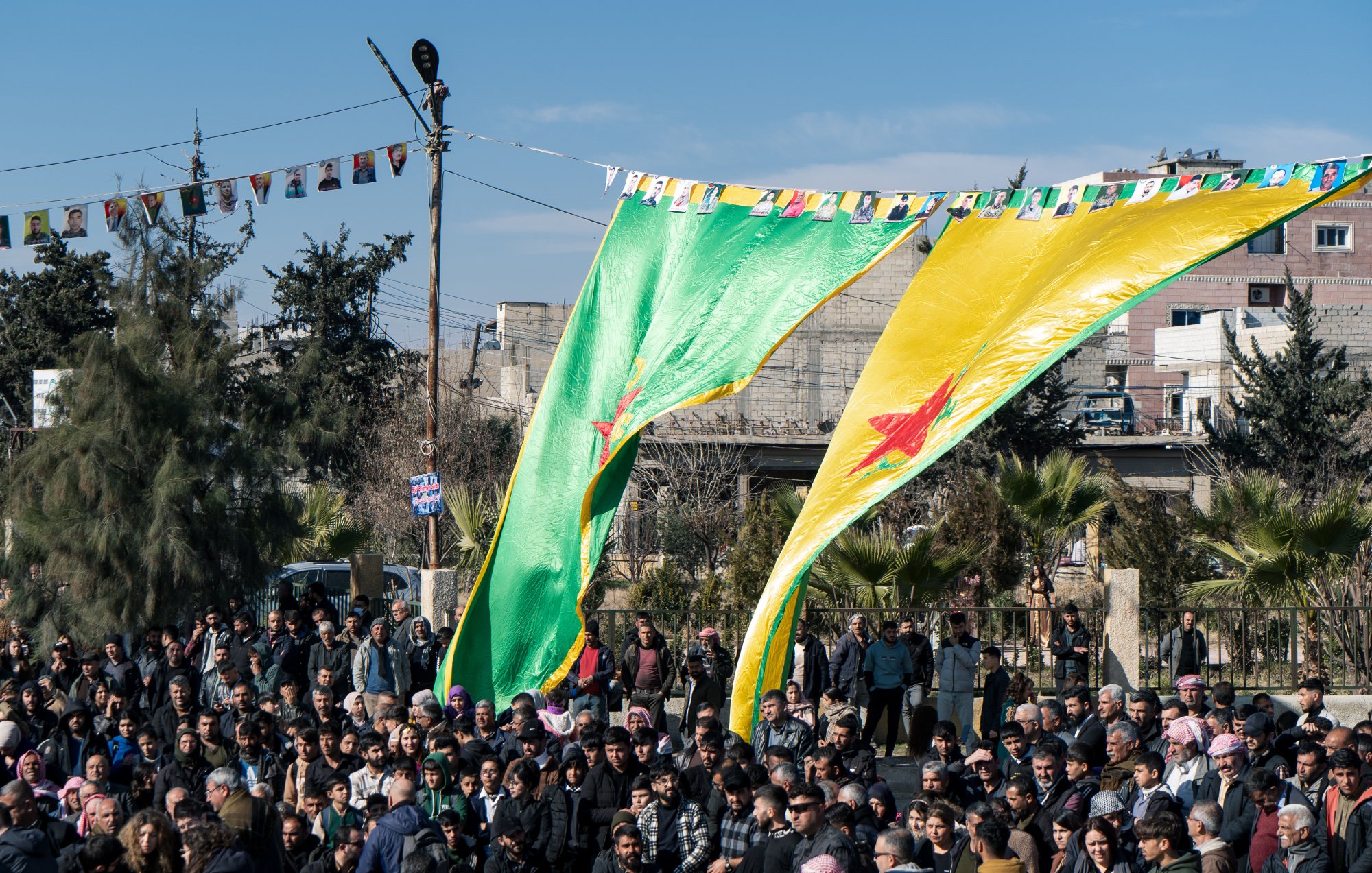 در این تصویر، تعدادی از افراد در یک تجمع یا مراسم حضور دارند. در پسزمینه، پرچمهای بزرگ با رنگهای سبز و زرد به همراه ستارهای قرمز به اهتزاز درآمدهاند. مردم در حال ایستادن و تماشا کردن هستند و درختان و ساختمانهایی در دوردست مشاهده میشود. همچنین، پرچمهای کوچکتری به نامهای مختلف در بالای محل تجمع آویزان شدهاند. آسمان آبی است و جو به نظر شاداب بهنظر میرسد.