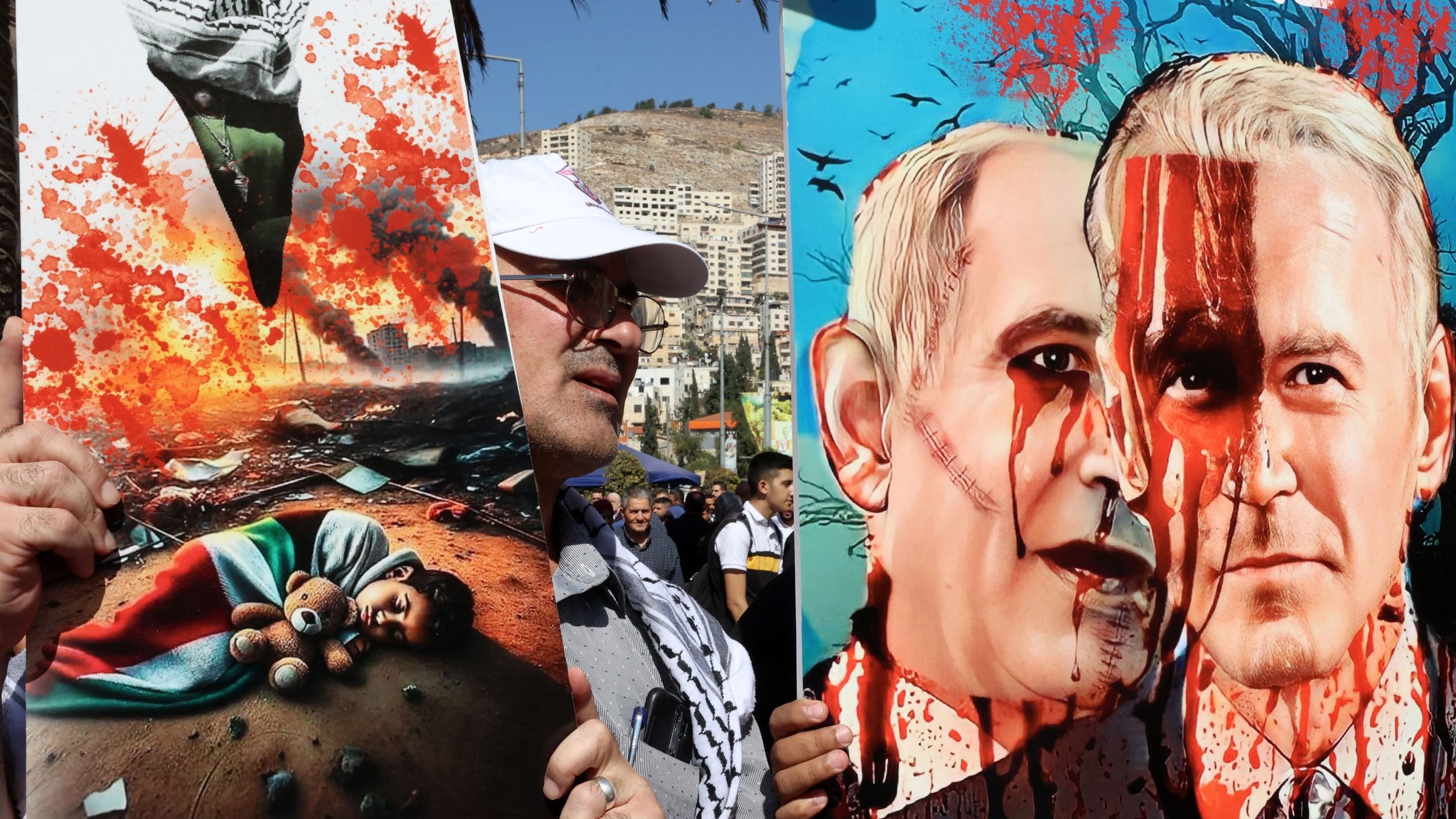 در این تصویر، دو تابلو بزرگ دیده میشوند که در مراسمی ممکن است مورد استفاده قرار گرفتهاند. تابلو سمت چپ تصویری غمانگیز از یک کودک در یک محیط ویران شده را نشان میدهد، با جزئیات مانند عروسکی در کنار کودک. تابلو سمت راست نیز چهرههای دو نفر را به تصویر میکشد که با خون آغشته شدهاند و نشاندهنده تنش یا اعتراض است. به نظر میرسد این تصاویر به موضوعات سیاسی و اجتماعی حساس اشاره دارند و بر احساسات شدید مربوط به آنها تأکید میکنند.