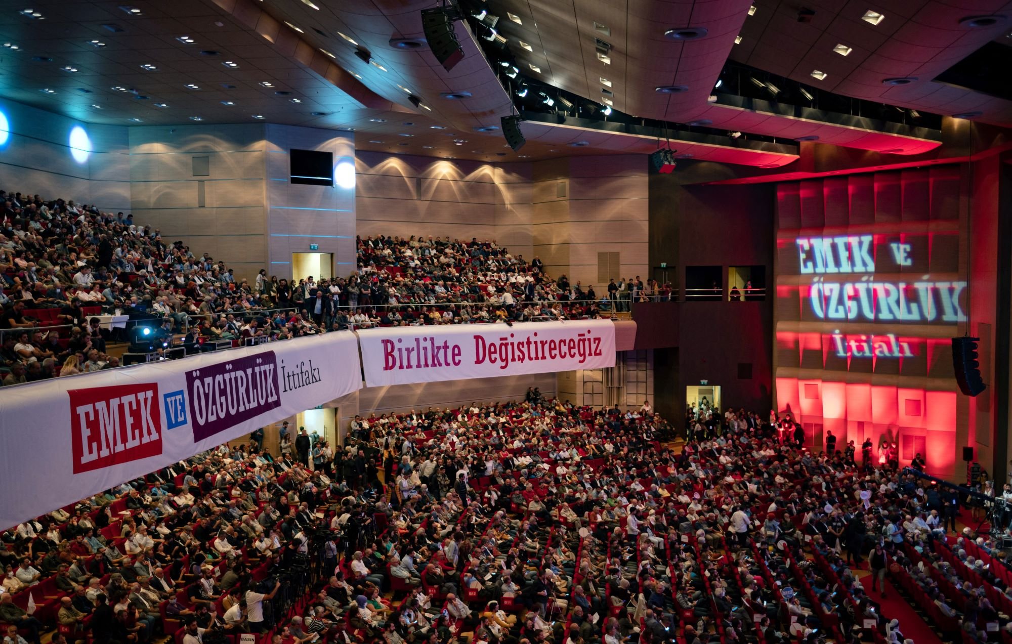 در این تصویر، صحنهی یک سالن بزرگ کنفرانس با تعدادی بسیار زیاد از افراد در حال نشستن و تماشا دیده میشود. سالن به خوبی روشن است و روی دیوارهای آن نوشتههایی به زبان ترکی وجود دارد. یکی از این نوشتهها به صورت برجسته "EMEK VE ÖZGÜRLÜK" (به معنی "کار و آزادی") است. فضا به نظر میرسد مربوط به یک همایش یا تجمع سیاسی باشد که در آن مردم به موضوعاتی مهم توجه دارند. در پسزمینه، نورپردازی خاصی وجود دارد که بر احساسات و فضای کنفرانس تأکید میکند.
