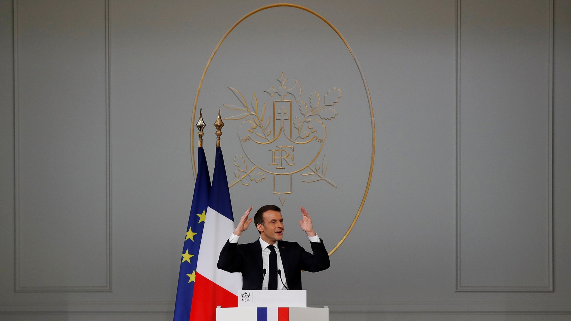 در این تصویر، یک مرد در حال سخنرانی است. او در جلوی یک تریبون ایستاده و دستهایش را به سمت بالا برده است. در پشت او، نماد رسمی فرانسه وجود دارد که شامل دو پرچم میباشد؛ یکی پرچم فرانسه و دیگری پرچم اروپایی. دیوار پشت او ساده و به رنگ خاکستری است و نماد جمهوری فرانسه در مرکز قرار دارد. این صحنه نشاندهنده یک رویداد رسمی یا کنفرانس مطبوعاتی است.