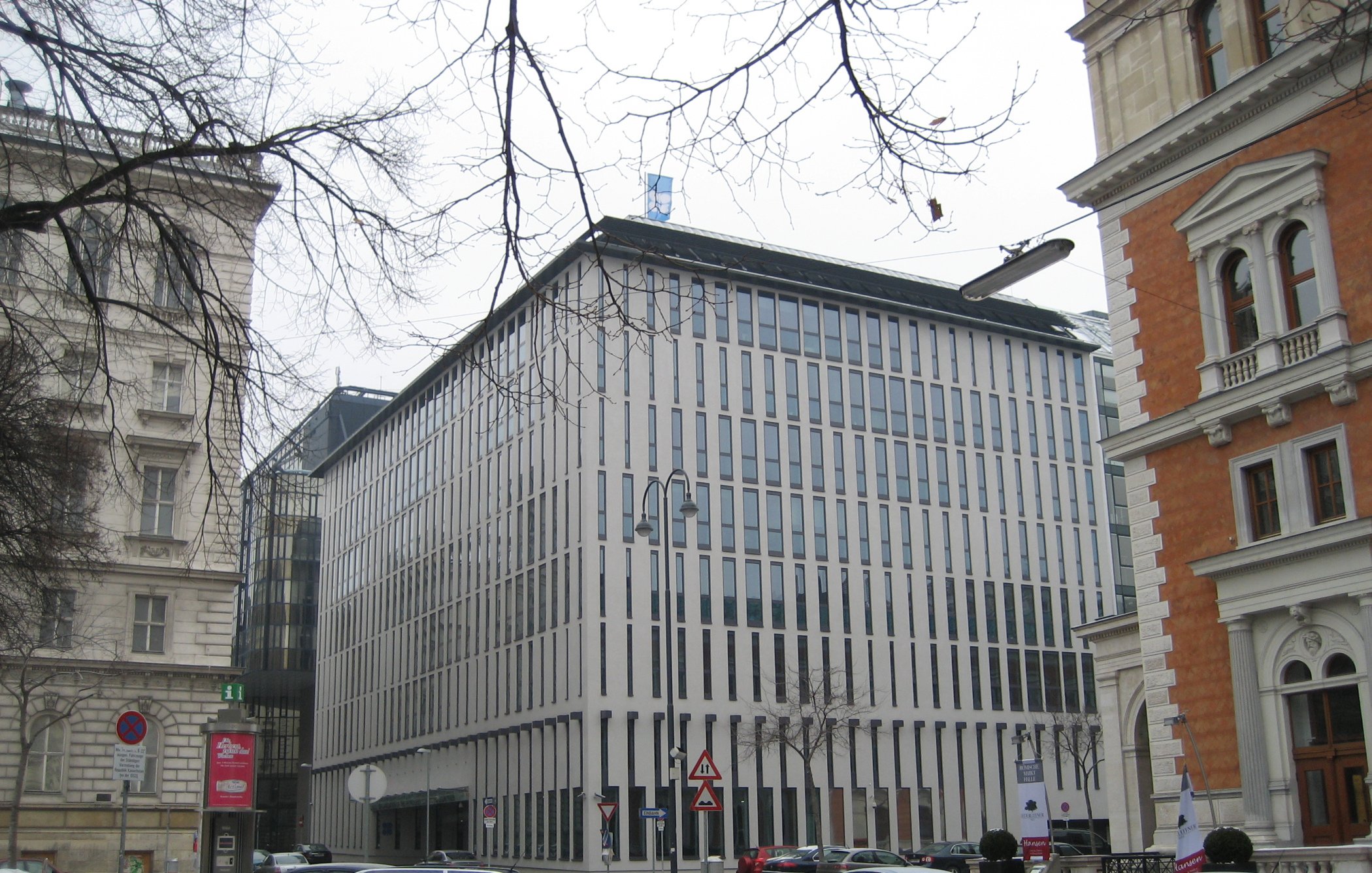 این تصویر نمای یک ساختمان مدرن را نشان میدهد که دارای طراحی مینیمالیستی است. نمای ساختمان با پنجرههای عمودی و خطوط باریک تقسیم شده، ظاهری مرتب و شیک دارد. اطراف ساختمان ممکن است شامل خودروها و درختان بدون برگ در فصل زمستان باشد. در سمت چپ و راست تصویر، ساختمانهای دیگری با طراحیهای کلاسیکتر وجود دارند که تضادی جالب با ساختمان مدرن ایجاد میکنند. فضا به نظر میرسد که در یک منطقه شهری و ممکن است در فصل سرد سال باشد.