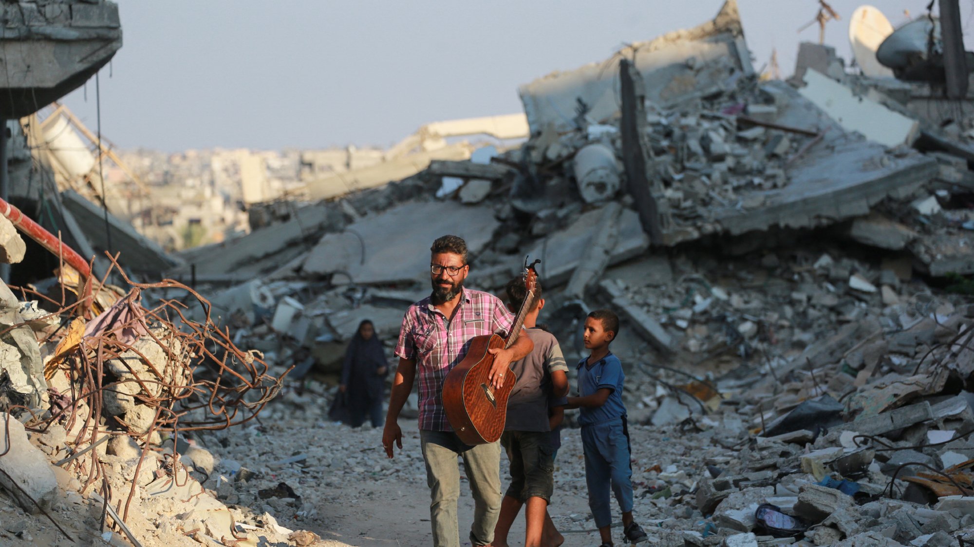 تصویر یک مرد را نمایش میدهد که در حال حرکت در میان ویرانههای یک منطقه تخریبشده است. او یک گیتار به دوش دارد و به نظر میرسد در حال نواختن یا آماده نواختن موسیقی باشد. دو کودک نیز به او نزدیک هستند، یکی از آنها به مرد وابسته است و به نظر میرسد او را دنبال میکند. در پسزمینه، آوار و خرابههای ساختمانها دیده میشود که نشانهای از یک بحران یا جنگ را نشان میدهد. نورخورشید در آسمان دیده میشود که ممکن است تصاویری از امید را القا کند.