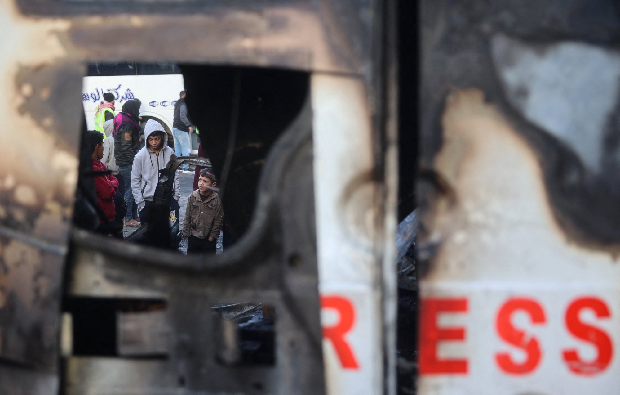 تصویر نشاندهندهی یک صحنهی غمانگیز است که در آن چندین کودک و بزرگسال درحال مشاهدهی یک وضعیت بحرانی به نظر میرسند. در پسزمینه، یک وسیله نقلیه آسیبدیده دیده میشود که حالت سوخته و خراب دارد. نام "پرس" بر روی آن به وضوح مشاهده میشود. در داخل نمای این وسیله، افرادی با حالتهایی جدی و نگران دیده میشوند که شاید به دنبال کمک یا راهی برای فرار از وضعیت خطرناک باشند. احساس تنش و اضطراب در این تصویر محسوس است.