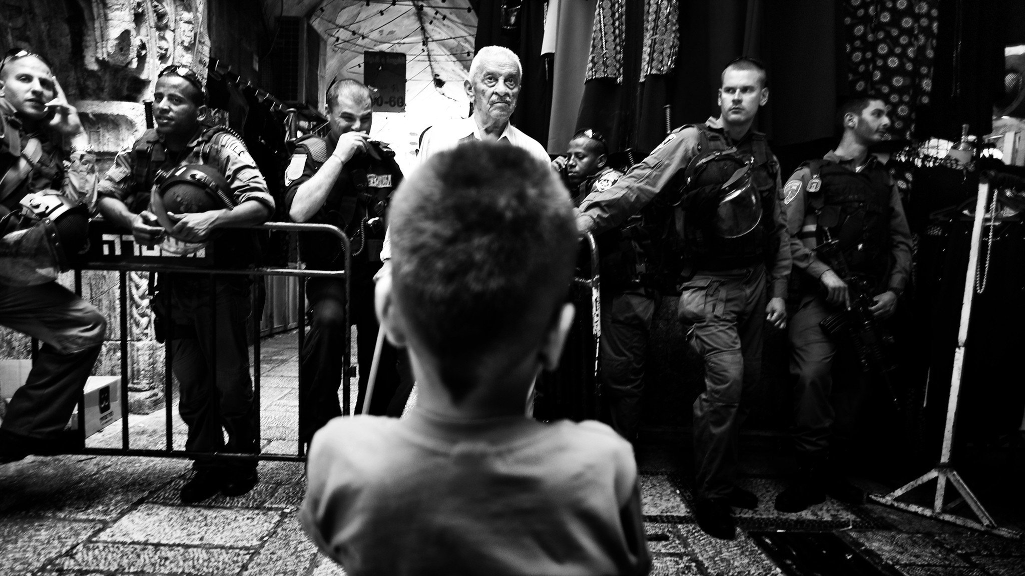 این تصویر سیاه و سفید، یک صحنه یاری را نشان میدهد که در آن یک کودک در حالت تماشا، به دو مرد بزرگسال و یک گروه از نیروهای امنیتی نگاه میکند. کودک از پشت تصویر دیده میشود و توجه او به مردی که در مقابلش ایستاده جلب شده است. نیروهای امنیتی در کنار آنها قرار دارند و به نظر میرسد که صحنهای از تنش یا تعامل میان افراد در حال رخ دادن است. فضای تصویر حس تنش و توجه را به خوبی منتقل میکند.