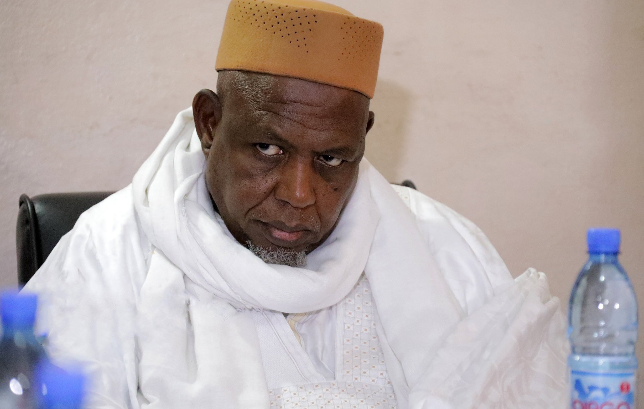 این تصویر یک مرد را نشان میدهد که لباسی سنتی به تن دارد و یک سرپوش قهوهای رنگ بر سر گذاشته است. او در حال نشسته و به جلو خیره شده است، به نظر میرسد که در حال تفکر یا گوش دادن به صحبتهای دیگران است. پسزمینه تصویری ساده و بدون هیچ جزئیات خاصی دارد.