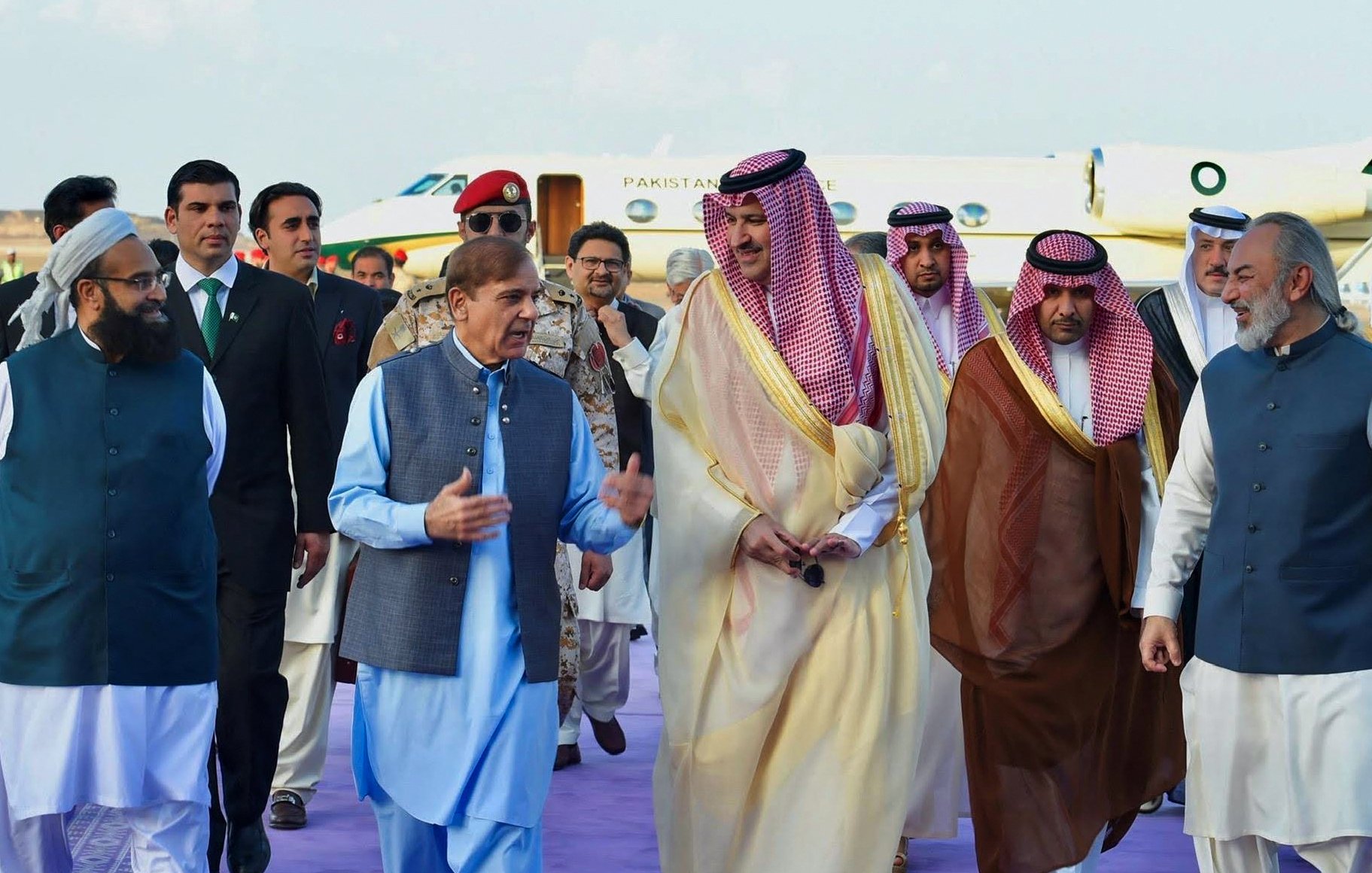 در این تصویر، گروهی از افراد در یک مراسم رسمی یا استقبال دیده میشوند. در مرکز تصویر، مردی با لباس سنتی پاکستانی در حال صحبت با دیگران است. اطراف او افرادی با لباسهای مختلف، از جمله لباسهای رسمی و سنتی، به چشم میخورند. پسزمینه شامل یک هواپیمای خصوصی است که نشاندهنده موقعیت ویژه این ملاقات است. فضا به نظر میرسد که محترمانه و دوستانه باشد.