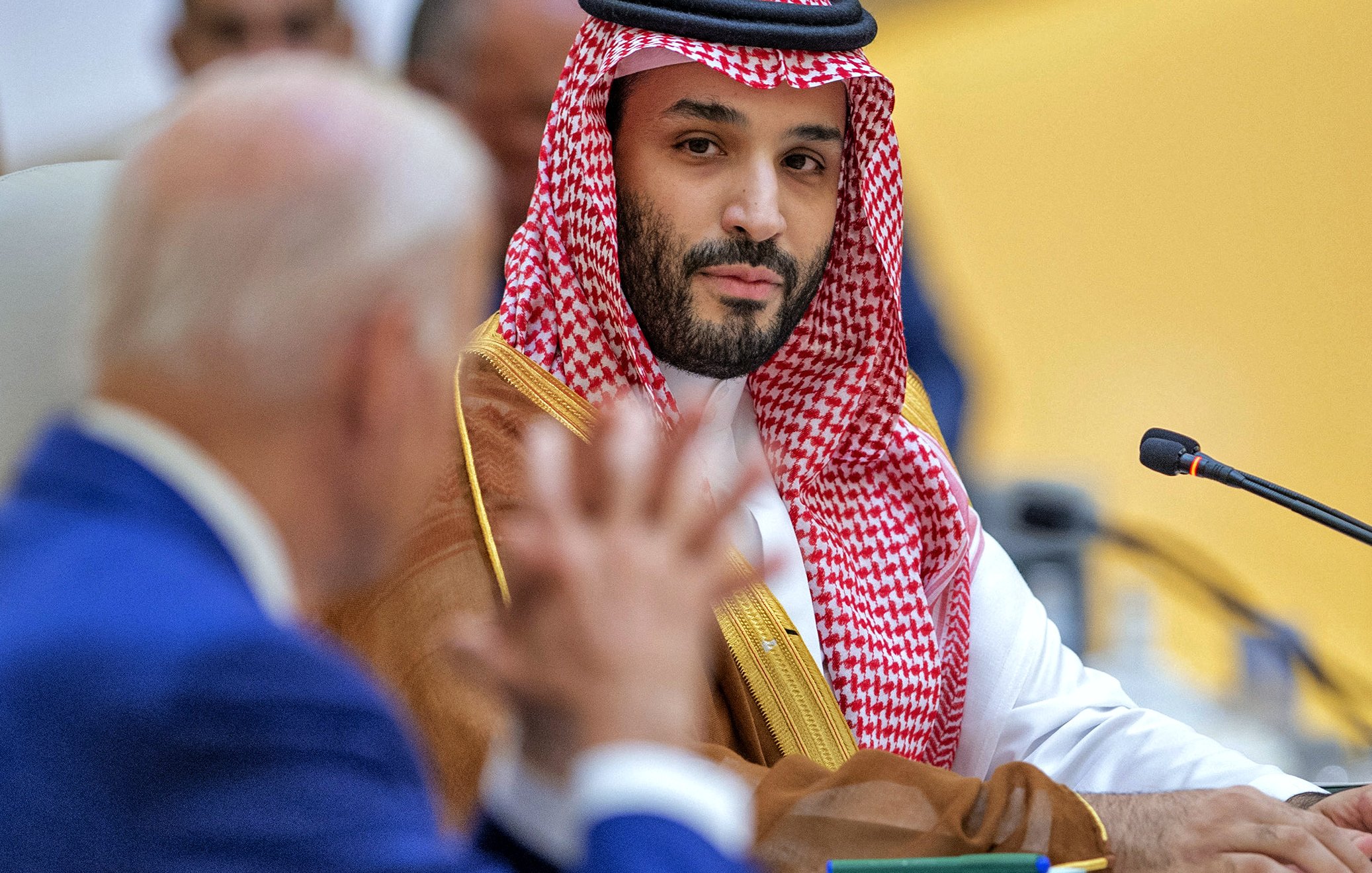 این تصویر مربوط به یک نشست یا جلسه رسمی است. در سمت چپ، یک مرد با لباس رسمی و کت و شلوار آبی نشسته است که به نظر میرسد در حال صحبت کردن است. در سمت راست، یک مرد دیگر با لباس سنتی عربی و عمامه قرمز و سفید، در حال گوش دادن به سخنان فرد مقابل است. پسزمینه سالن جلسه به طرزی طراحی شده که نشاندهنده یک محیط رسمی و دیپلماتیک است.