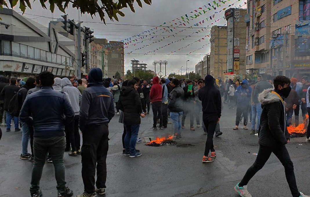 تصویر نشاندهنده یک تجمع اعتراضی در یک خیابان است. در پسزمینه، تعدادی از ساختمانها دیده میشود و آسمان به رنگ خاکستری است. جمعیت زیادی از افراد در لباسهای گرم در حال ایستادن یا حرکت کردن هستند. برخی از آنها ماسک به چهره دارند. در چند نقطه از خیابان، دود و شعلههایی به چشم میخورد که نشاندهنده وجود آتش است. همچنین، تزئینات و پرچمهایی در بالای خیابان دیده میشود که به نظر میرسد نشاندهنده یک مناسبت خاص یا جشنواره است. جمعیت به نظر میرسد که در حال تظاهرات یا اعتراض هستند.