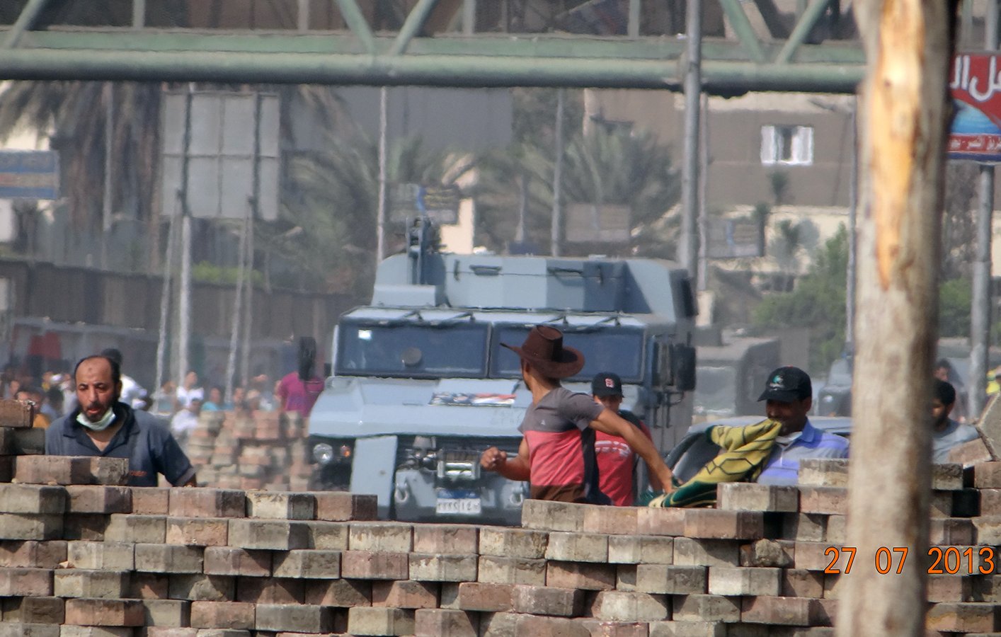 در این تصویر، تعدادی کارگر در پشت دیواری از آجرهای شکسته در حال کار هستند. در پسزمینه، یک خودروی نظامی یا زرهی با ظاهر مشکی دیده میشود که به سمت آنها نزدیک میشود. فضای شهر مشخص است و تعدادی درخت در پسزمینه وجود دارد. تاریخ تصویر نیز در گوشه پایین سمت راست قرار دارد که نشان میدهد این عکس در تاریخ ۲۷ ژوئیه ۲۰۱۳ گرفته شده است. جو کلی تصویر به نظر میرسد در حالتی تنشزاست.