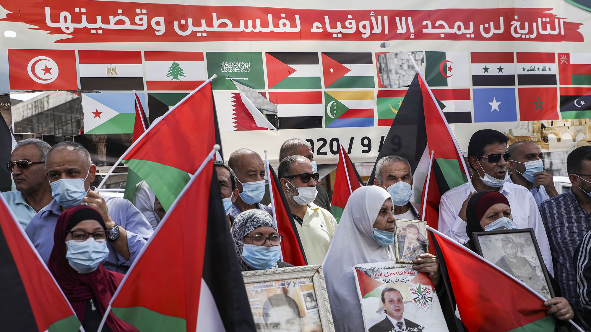 در این تصویر، گروهی از مردم در حال برگزاری یک تجمع هستند. آنها پرچمهای فلسطین را در دست دارند و برخی از آنها عکسهایی از شخصیتهای مهم را حمل میکنند. بر روی پسزمینه، پرچمهای کشورهای مختلف دیده میشود که نشان از حمایتهای بینالمللی از قضیه فلسطین دارد. همچنین، بسیاری از شرکتکنندگان دارای ماسک هستند، که ممکن است به دلیل رعایت مسائل بهداشتی باشد. احساس همبستگی و حمایت از آرمان فلسطین در چهرههای آنها نمایان است.