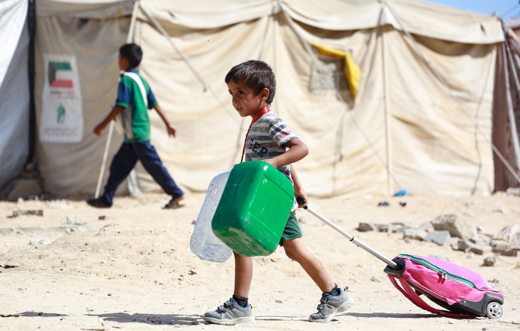 در تصویر، پسربچهای دیده میشود که در حال حمل یک کولهپشتی به رنگ صورتی و یک ظرف سبز رنگ است. او در یک محیط بیابانی و خشک قرار دارد که چادرهای موقتی در پسزمینه به چشم میخورد. پسربچه لباسی ساده و راحت پوشیده و به نظر میرسد که در حال حرکت به سمت مقصدی است. در کنار او، کودک دیگری نیز در حال گذر است. فضای تصویر حالتی از زندگی در شرایط سخت را به نمایش میگذارد.