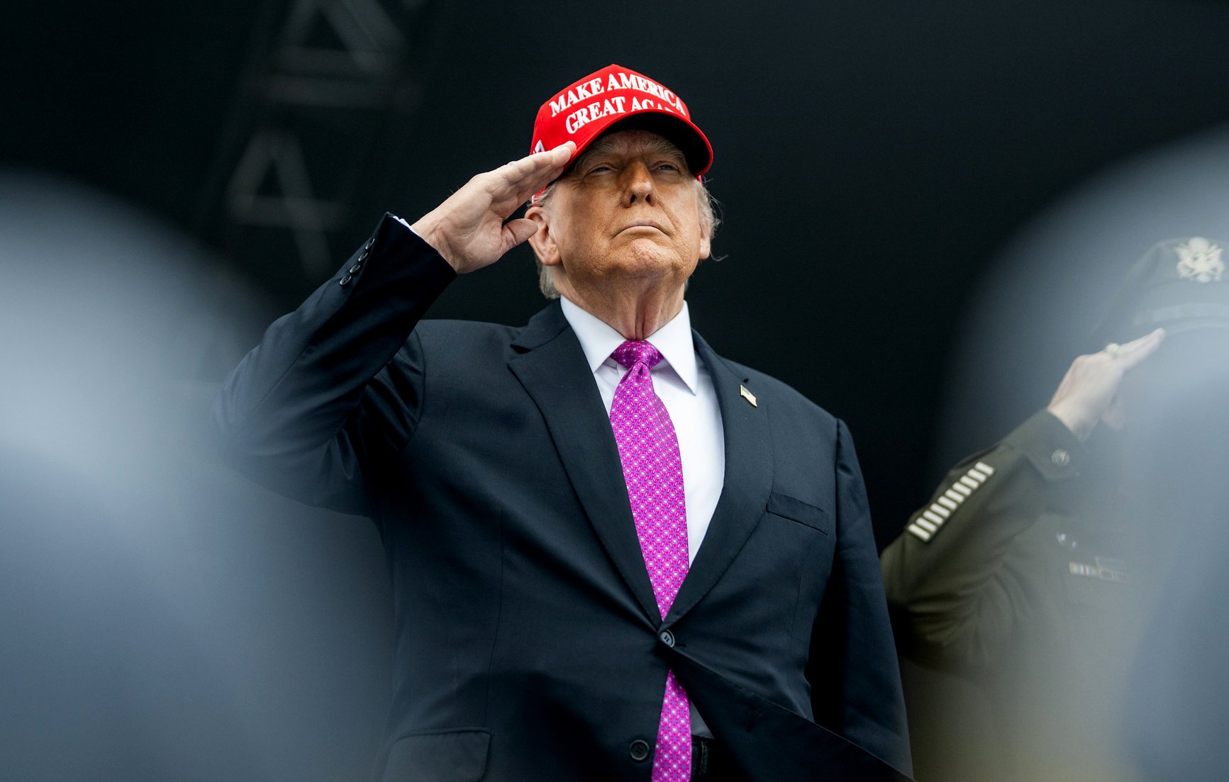 مردی با کلاهی قرمز و پیراهن مشکی، با دست بر پیشانی ایستاده و سلام میکند.