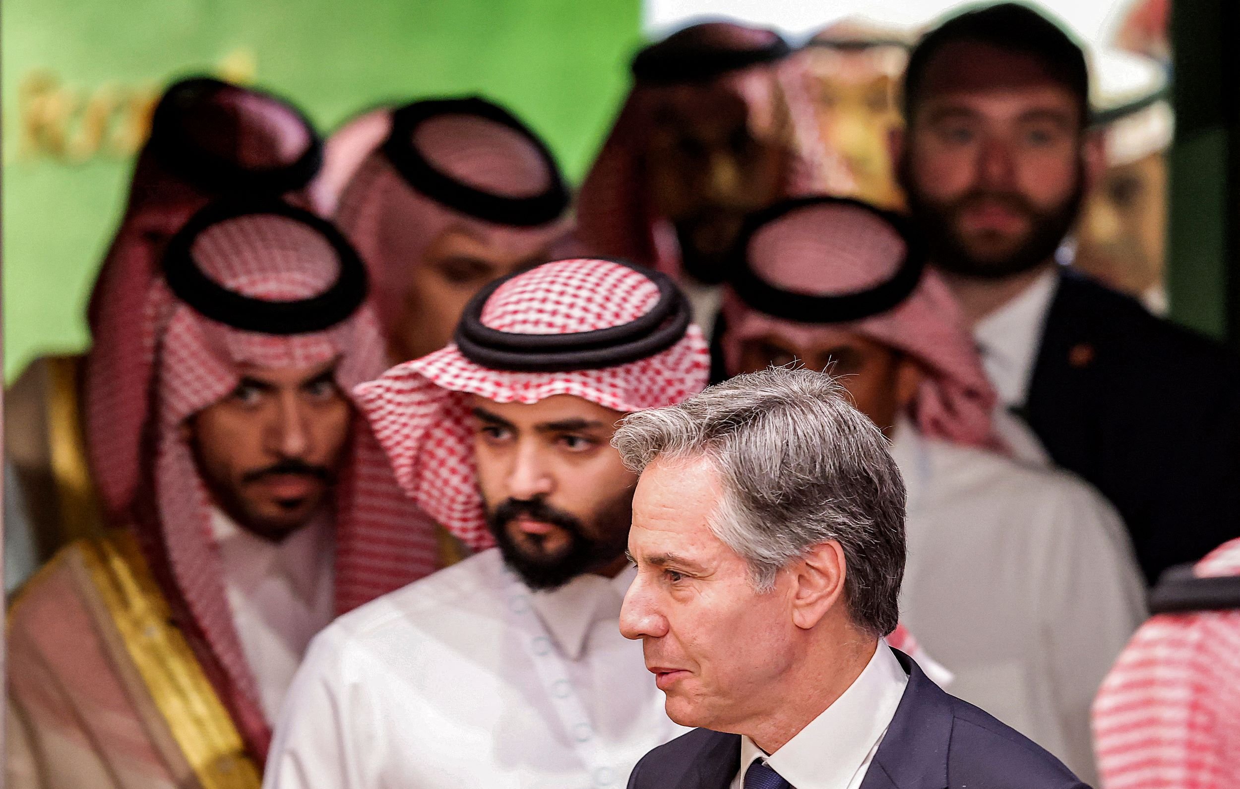 در این تصویر، تعدادی مرد که به نظر میرسد لباسهای سنتی عربی پوشیدهاند، در پسزمینهای روشن و زنده قرار دارند. در جلو، مردی با کت و کراوات به سمت راست در حال حرکت است. به نظر میرسد این تصویر مربوط به یک نشست یا مراسم رسمی باشد. فضای کلی تصویر حس تعامل و گفتوگو را منتقل میکند.