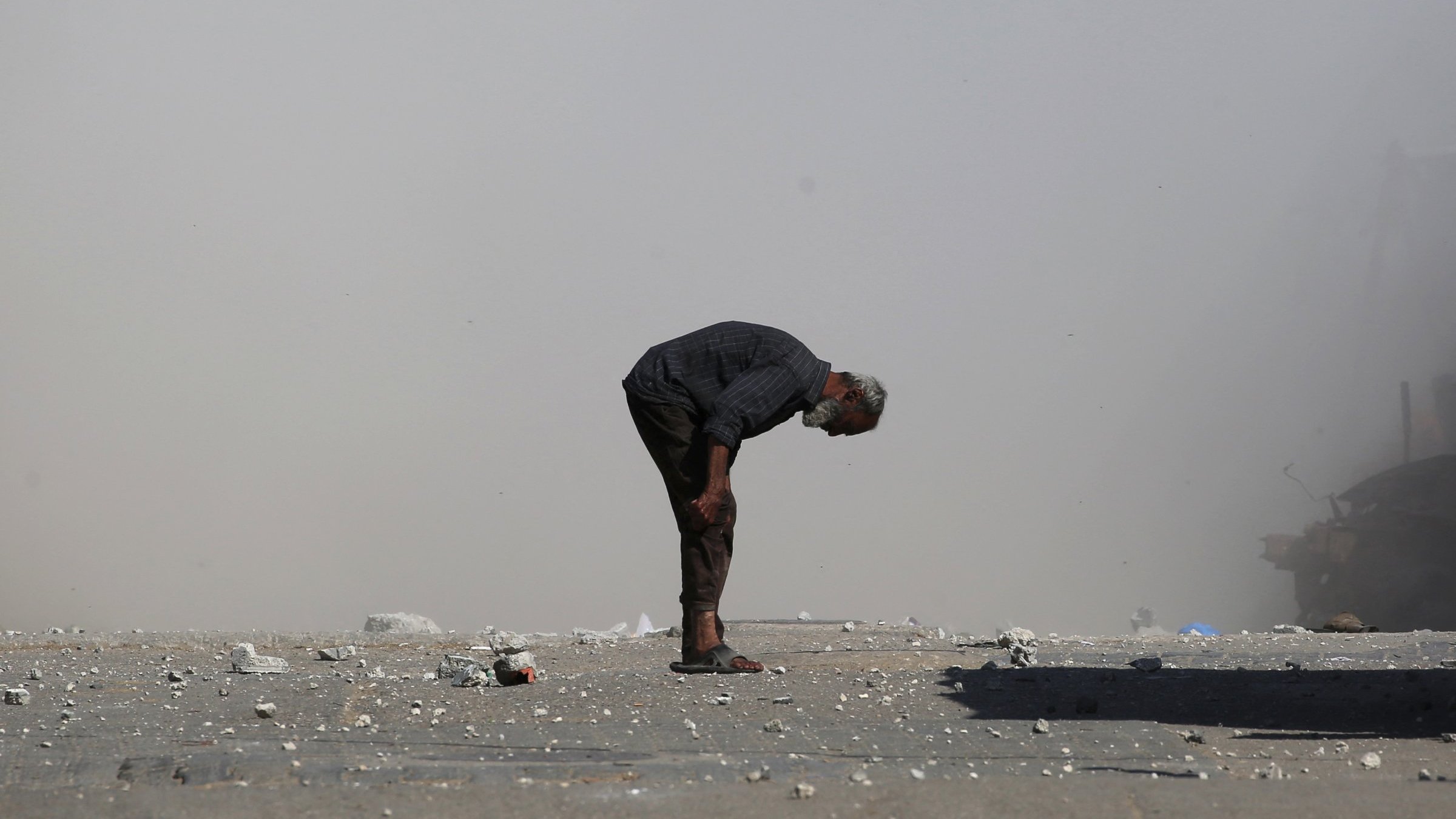 مردی مسن با کمر خمیده در مکانی خاکی و غبارآلود ایستاده است. اطرافش صحنهای ویران وجود دارد.