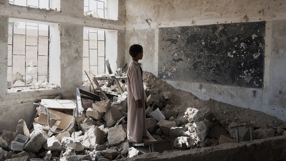 در این تصویر، یک پسر جوان در یک کلاس درس ویران شده ایستاده است. دیوارها و سقف کلاس آسیب دیدهاند و بخشهایی از زیرساختها به طور کامل تخریب شدهاند. در اطراف او، زبالهها و آوارهای ساختمانی وجود دارد. یک تابلو سیاه با نوشتههایی بر روی دیوار وجود دارد، اما وضعیت آن نیز نامناسب است. نور از پنجرههای شکسته به فضای داخلی میتابد و حس ناامیدی و ویرانی را منتقل میکند.