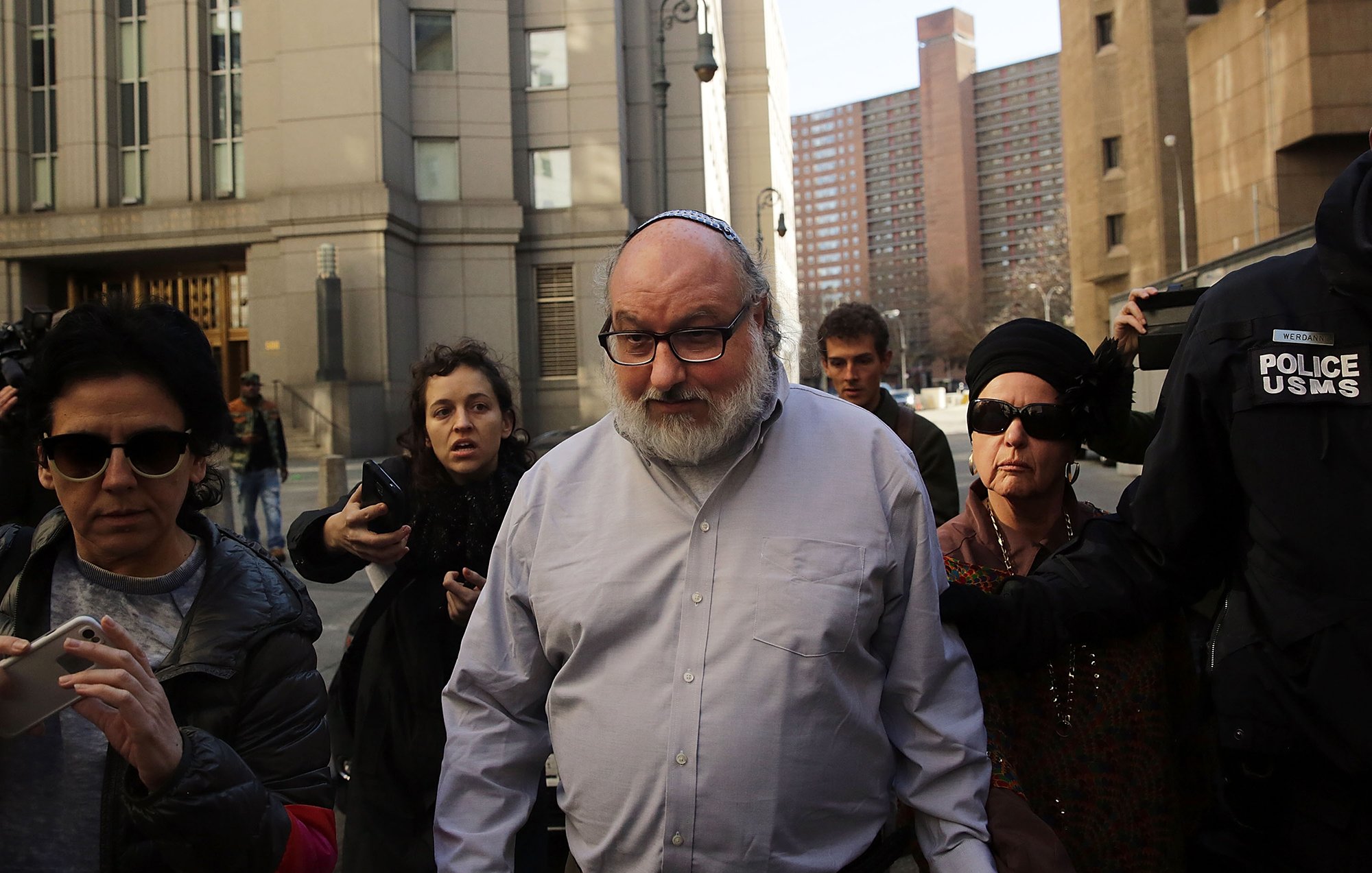 در این تصویر، یک مرد میانسال با عینک و کلاه کیپا در حال خروج از یک ساختمان به نظر میرسد. او به سمت دوربینها و گروهی از خبرنگاران توجه دارد. چند زن هم در اطراف او هستند که یکی از آنها عینک دودی به چشم دارد و دیگری کلاهی بر سر دارد. محیط اطراف شهری به نظر میرسد و چندین ساختمان بلند در پسزمینه قابل مشاهده است. جو تصویر حس هیجان و توجه رسانهها را منتقل میکند.