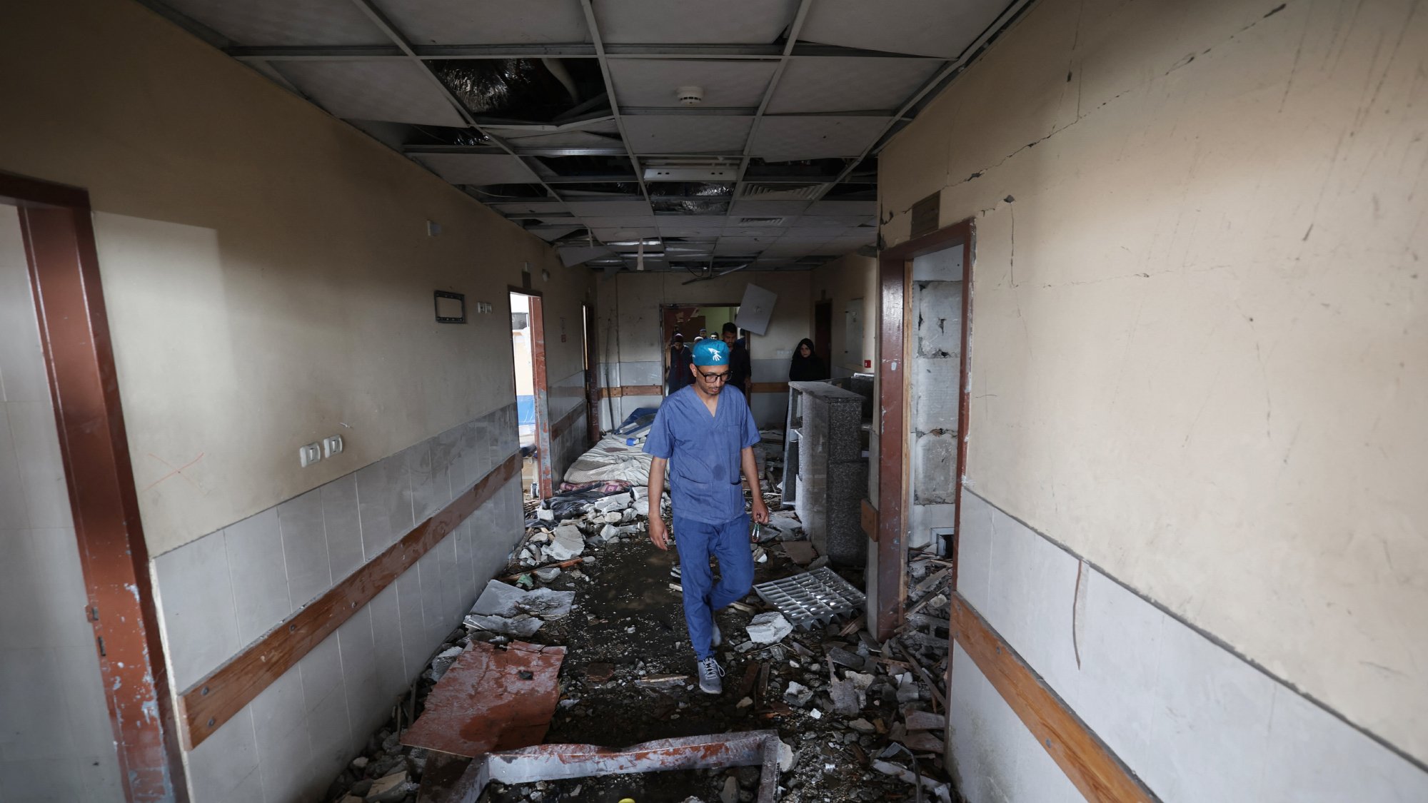 مردی با کلاه ایمنی در راهروی ویران یک بیمارستان قدم میزند، دیوارها آسیب دیده و کف پر از آوار است.
