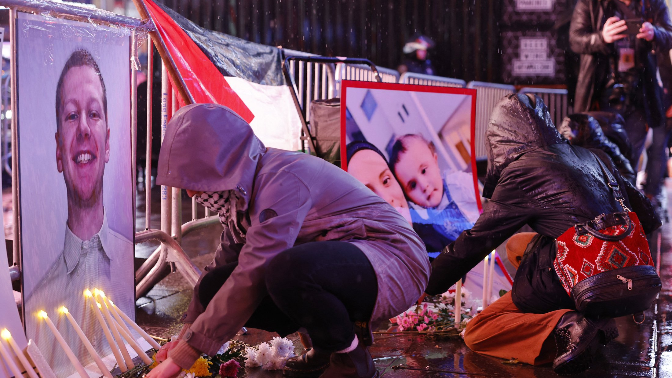 تصویر فضای یک تجمع یادبود را نشان میدهد که در آن افراد در حال قرار دادن شمع و گل هستند. در جلوی آنها، تصاویری از یک فرد و یک خانواده به نمایش گذاشته شده است. باران به زمین میریزد و خیابان براق است. افراد با لباسهای گرم و کلاه روی زمین نشستهاند و به یاد افرادی که از دست رفتهاند، مراسمی برگزار میکنند. این صحنه حال و هوای غم و اندوه را منتقل میکند.