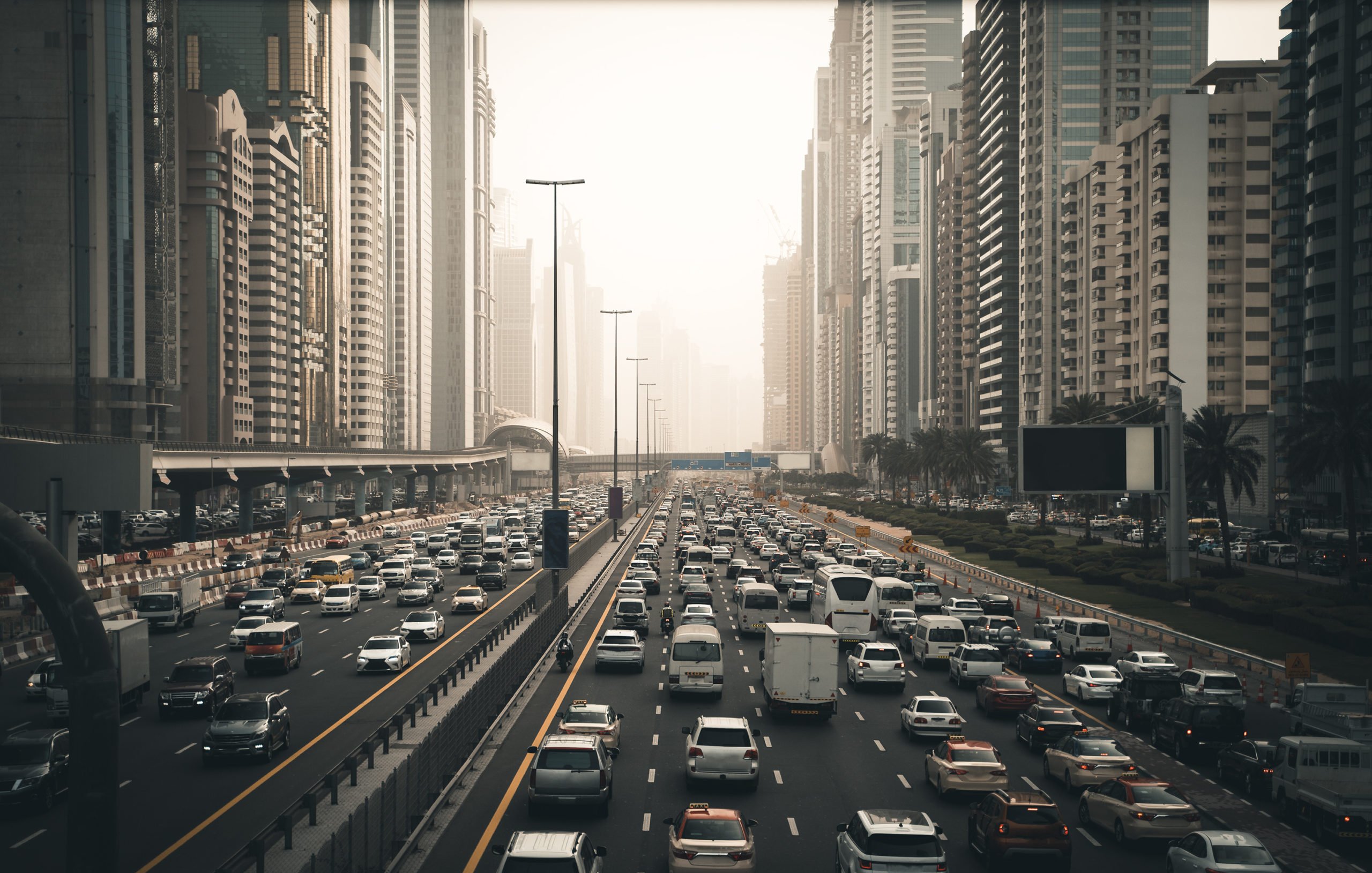 این تصویر نمایی از یک خیابان شلوغ در یک شهر مدرن را نشان میدهد. در سمت چپ، ساختمانهای بلند و مرتفع با معماریهای معاصر دیده میشوند. خیابان مملو از خودروهاست که در دو طرف حرکت میکنند. ترافیک به وضوح قابل مشاهده است و برخی خودروها در حالت توقف به نظر میرسند. در زمینه، مه یا گرد و غباری به چشم میخورد که باعث کاهش وضوح چشمانداز میشود. فضای کلی تصویر حس شلوغی و پویا بودن شهر را منتقل میکند.
