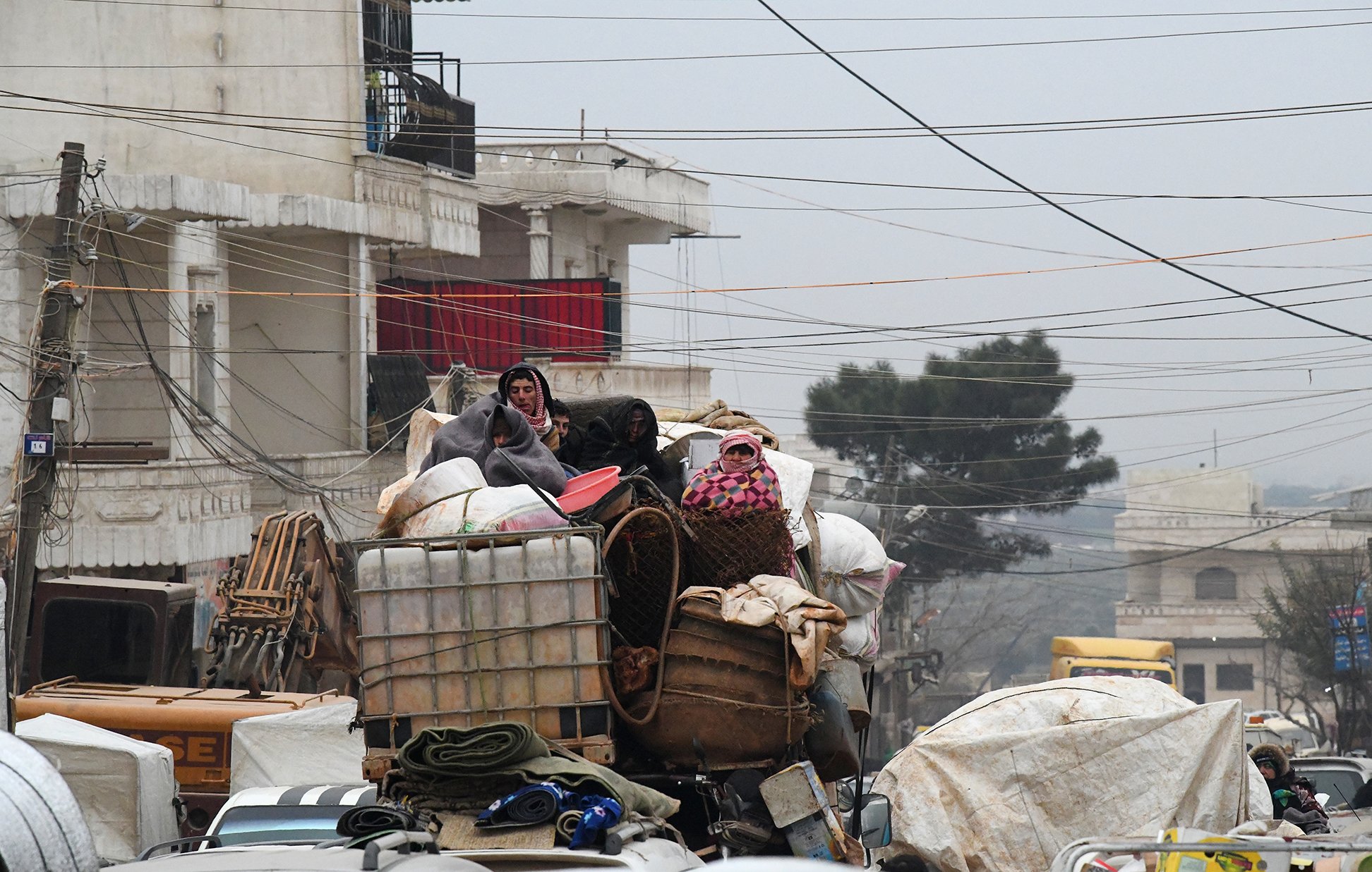 در این تصویر، گروهی از افراد در حال سفر بر روی یک کامیون بار هستند. آنها به طور واضحی در میان بار زیادی از محصولات و وسایل قرار دارند. برخی از افراد پوشیده از لباسهای ضخیم و شالهای رنگی به نظر میرسند که به دلیل سرما به خود پیچیدهاند. محیط اطراف شلوغ و شهرنشین به نظر میرسد، با ساختمانها و خطوط برق مشخص. وضعیت کلی تصویر احساس چالش و سختی را القا میکند.