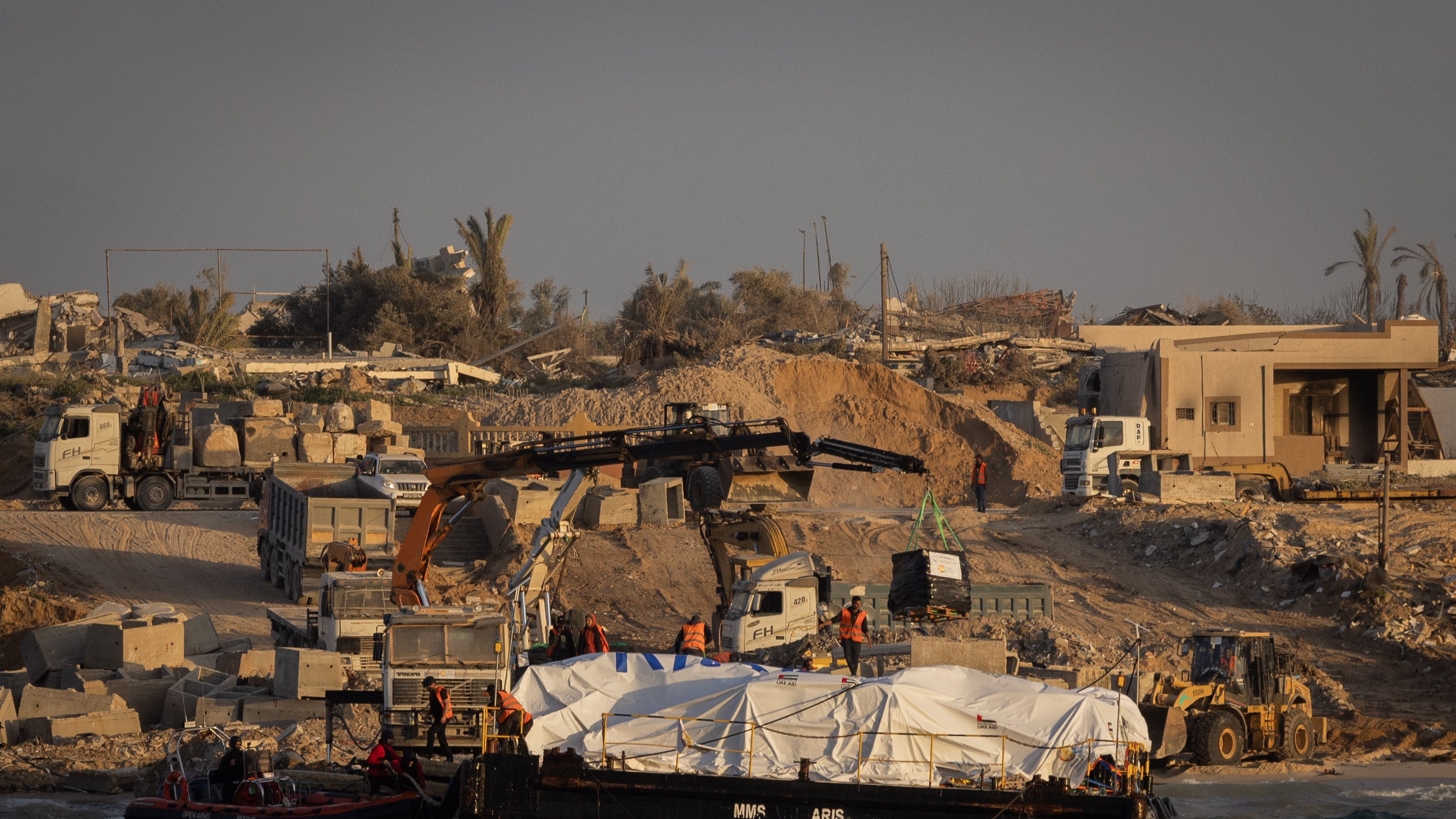 در این تصویر، یک صحنه ساختمانی در کنار دریا دیده میشود. در ساحل، تعدادی ماشین سنگین و جرثقیل در حال کار هستند. یک قایق روی آب قرار دارد که زیر آن یک پوشش سفید رنگ قرار دارد. پسزمینه شامل ساختمانهای نیمهکار و تخریب شده و چند درخت نخل است. نور خورشید به صورت گرم بر روی این صحنه تابیده و حال و هوای آن را به تصویر کشیده است.
