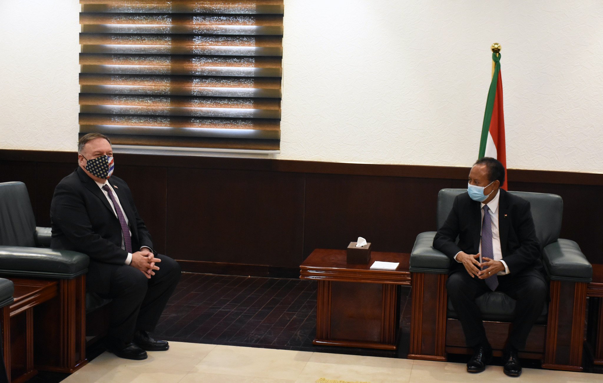 در این تصویر، دو مرد در یک اتاق رسمی نشستهاند. یکی از آنها که سمت چپ تصویر است، ماسک دارد و ظاهری رسمی با کت و شلوار پوشیده است. مرد دیگر در سمت راست نیز لباس رسمی به تن دارد و به طرف مرد اول نگاه میکند. در پسزمینه، پردههای پنجره و پرچم یک کشور مشخص است. فضا به وضوح رسمی و موقر به نظر میرسد و به احتمال زیاد مربوط به یک ملاقات دیپلماتیک است.