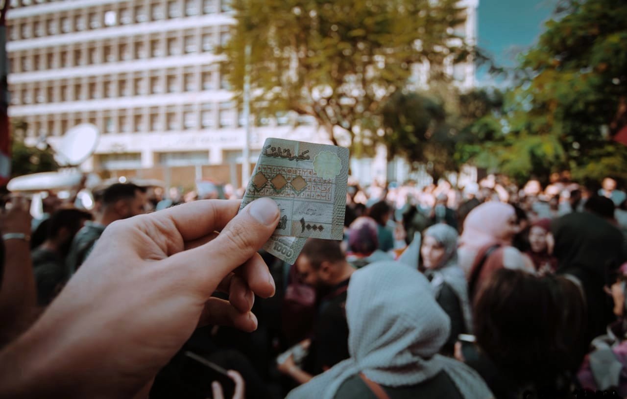 در این تصویر، یک دست در حال نگهداشتن یک کارت شناسایی یا مدرک مشابهی است. در پسزمینه، جمعیتی از افراد دیده میشود که در یک مکان عمومی جمع شدهاند. برخی از افراد چادر یا پوششهای اسلامی دارند. نور روز و فضای خارجی نشاندهنده یک رویداد یا تجمع عمومی است. احساس عمومی شلوغی و فعالیت اجتماعی در این تصویر مشهود است.