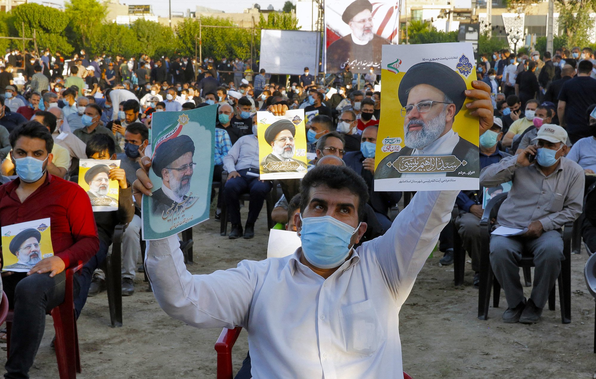 در تصویر، جمعیتی از افراد در یک فضای باز دیده میشود که روی صندلی نشستهاند و تعدادی از آنها در حال نگهداشتن پوسترهایی از یک شخصیت سیاسی هستند. مردم ماسک بهصورت دارند و در پسزمینه، تعدادی پرچم و تابلوهای نمایشی نیز وجود دارد. این جمعیت به نظر میرسد که در یک رویداد یا حمایت سیاسی شرکت کردهاند.