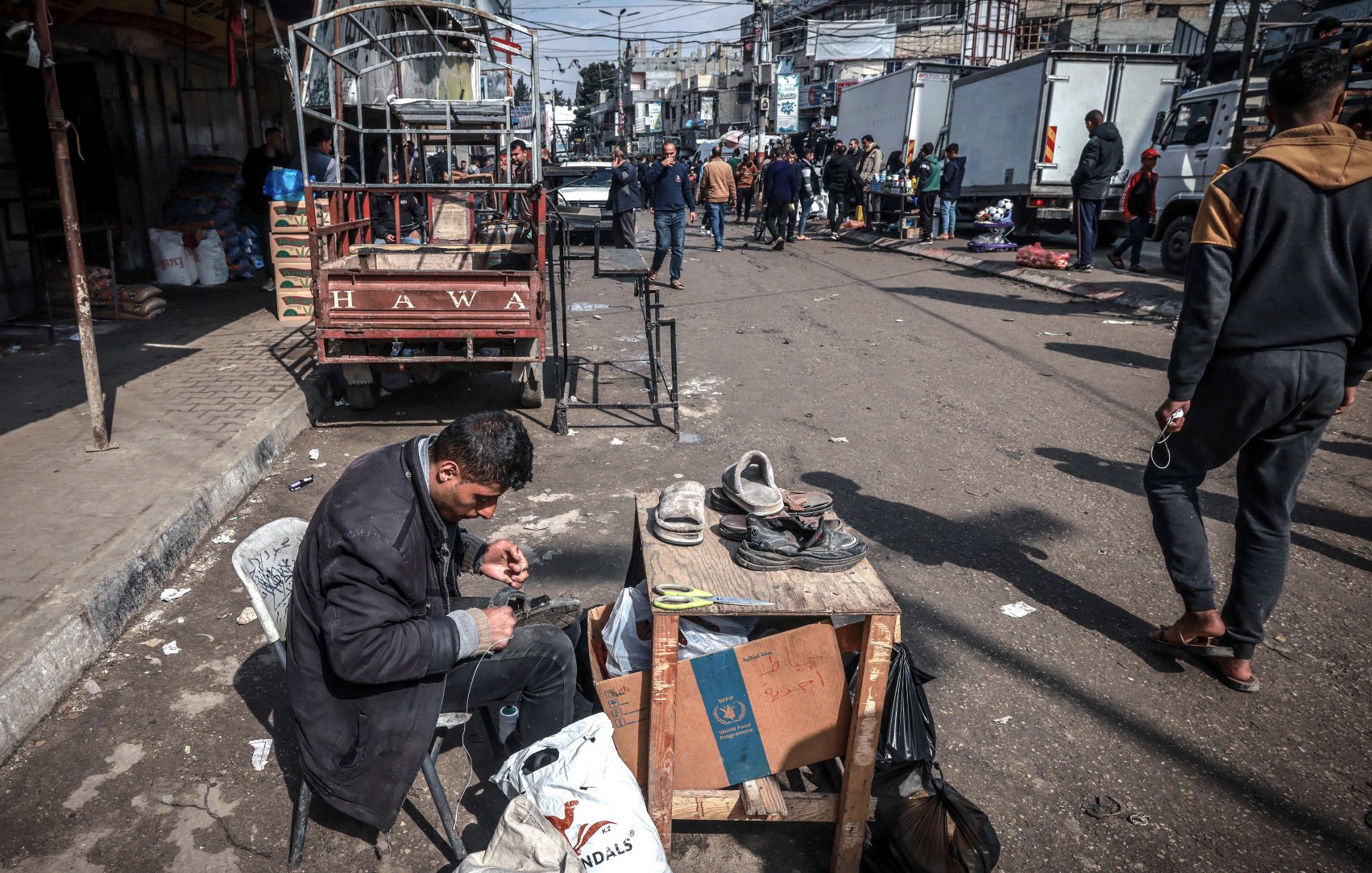 در تصویر، مردی در حال تعمیر یا واکس زدن کفشها است. او روی یک صندلی نشسته و وسایل کارش را بر روی یک میز کوچک چوبی قرار داده است. در اطراف او، افرادی در حال عبور و مرور هستند و تعدادی خودروی باربری در پسزمینه دیده میشود. فضایی شلوغ و شهری به نظر میرسد و فعالیتهای روزمره در حال انجام است. چند کشیده و بقیه اجناس نیز در اطراف قرار دارند.
