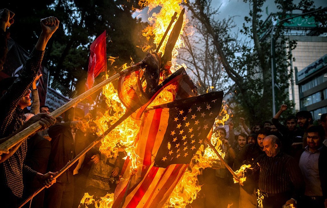 این تصویر نشاندهنده یک تجمع اعتراضی است که در آن گروهی از افراد در حال سوزاندن پرچم ایالات متحده هستند. در پسزمینه، دیده میشود که برخی از افراد دستها را بالا برده و به نشانه اعتراض فریاد میزنند. دود و آتش در حال برخاستن از پرچم است و فضا به شدت احساسی و پر از شور و هیجان به نظر میرسد. درختان و ساختمانهایی نیز در پسزمینه وجود دارند که نشاندهنده مکان شهری این رویداد است.
