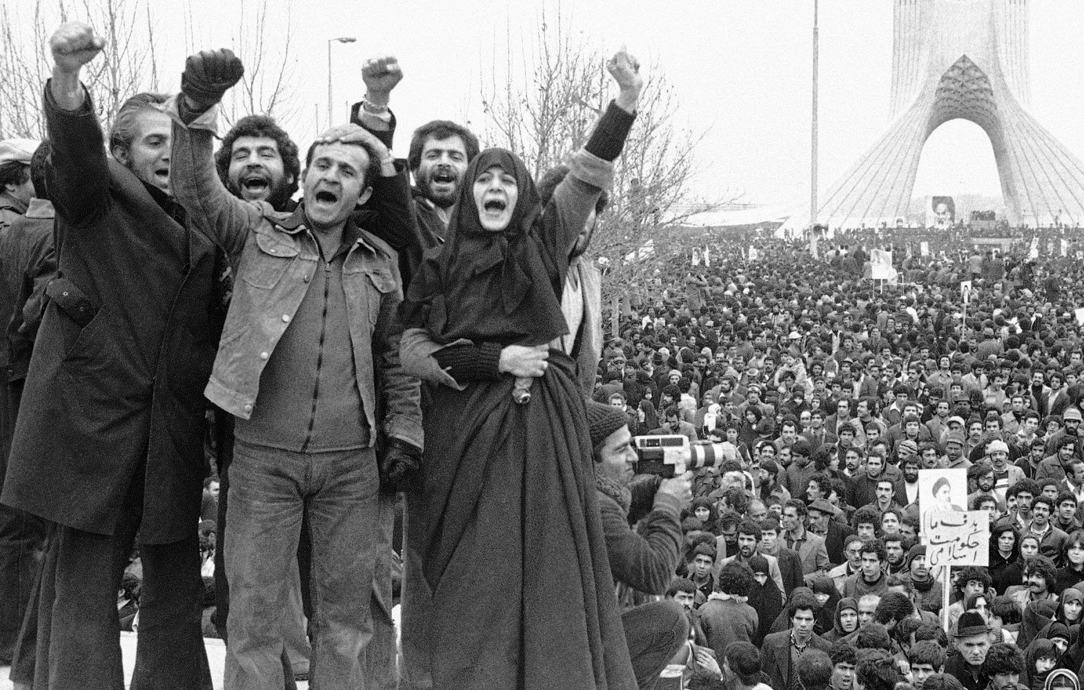 تصویر نمایی از یک تجمع بزرگ مردمی است که در میدان آزادی تهران برگزار شده است. در جلو، گروهی از افراد دستهای خود را بالا برده و به نشانه اعتراض یا ابراز شادی فریاد میزنند. آنها لباسهای متفاوتی بر تن دارند و برخی از آنها چهرههایشان را خوشحال یا هیجانزده نشان میدهند. در پسزمینه، جمعیت زیادی دیده میشود که به سمت دوربین و افراد جلویی نگاه میکنند. بنای میدان آزادی نیز در پسزمینه قابل مشاهده است و جوّ کلی تصویر نشاندهنده شور و هیجان یا اعتراض اجتماعی است.