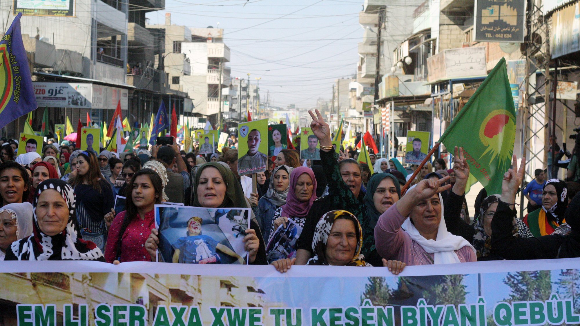 تصویر یک تظاهرات بزرگ را نشان میدهد که گروهی از زنان در آن شرکت دارند. آنها در خیابانی شلوغ راه میروند و پلاکاردهایی به دست دارند. تعدادی پرچم با نمادهایی خاص نیز در دست دارند. زنان با لبخند و دستهای بالا به نشانه حمایت و ابراز احساسات مشارکت میکنند. محیط اطراف شامل ساختمانها و جمعیت دیگری است که در پسزمینه دیده میشود. این تصویر به وضوح نشاندهنده یک حرکت اجتماعی یا سیاسی است.