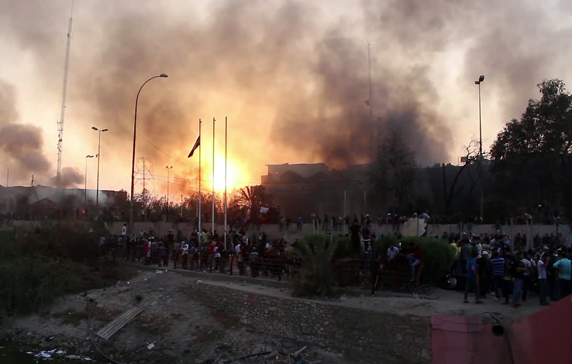 این تصویر نمایی از یک سناریو پرتنش را به تصویر میکشد، که شامل آتش و دود در آسمان است. غلظت دود نشاندهنده یک حادثه یا اعتراض شدید است. افرادی در پایین تصویر تجمع کردهاند و به نظر میرسد که در حال تماشا یا شرکت در یک رویداد مهم هستند. در پسزمینه، ساختمانهایی دیده میشود که به همراه تیرکهای پرچم و تجهیزات دیگر قابل توجه است. این صحنه به طور کلی حس ناامنی و هیجان را منتقل میکند.