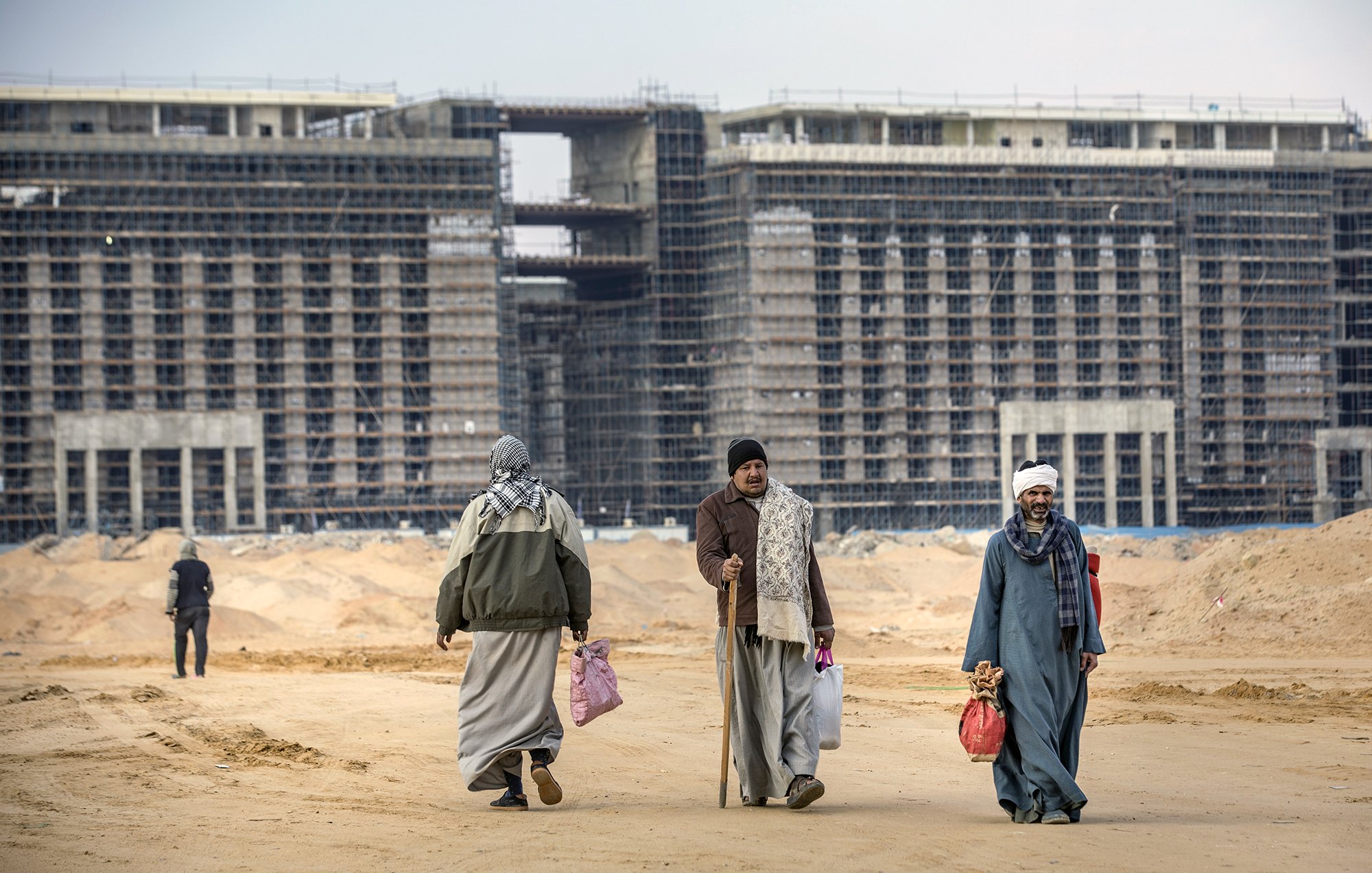 در تصویر، گروهی از مردان در محیطی بیابانی و ساختمانی در حال حرکت هستند. پشت سر آنها، ساختمانهای در حال ساخت دیده میشود که نمایی خالی و ناتمام دارند. افراد با لباسهای سنتی، برخی با چوبدست و کیسههایی در دست، در حال رفت و آمد در جادهای شنزاری هستند. آسمان کمی ابری به نظر میرسد و فضایی نسبتاً خنک و خاموش برقرار است.