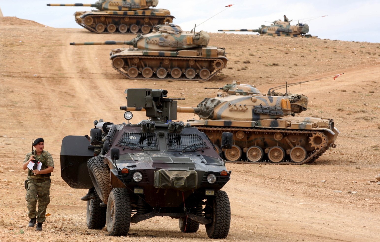 تصویر نشاندهنده یک صحنه نظامی است که در آن چندین تانک در حال حرکت یا منتظر هستند. در جلوی تصویر، یک خودرو نظامی سنگین قرار دارد که یک سرباز در حال بررسی یا نگهبانی در کنار آن ایستاده است. زمین کوهستانی و خشک به نظر میرسد و فضای کلی تصویر حس نظامی و جدی بودن را منتقل میکند. تانکها به سمت دورتر تصویر حرکت کرده و در حال آمادهباش به نظر میرسند.