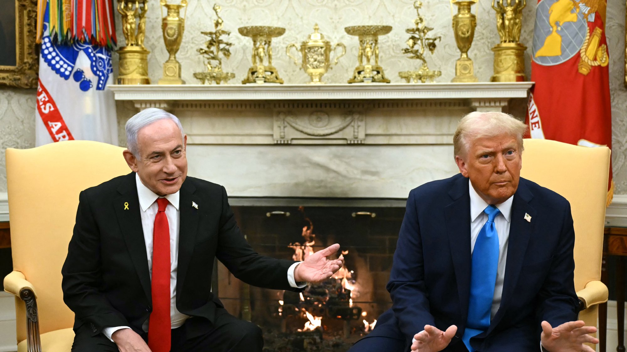 در این تصویر، دو شخصیت سیاسی در یک اتاق ملاقات نشستهاند. یکی از آنها در لباس رسمی سیاه و با یقه سفید و کراوات قرمز حاضر شده است و دیگری در کت و شلوار تیره و با کراوات آبی نشسته است. پسزمینه شامل یک شومینه و دکوراسیون کلاسیک است. هر دو شخصیت در حال گفتوگو به نظر میرسند و حرکات دست آنان نشاندهنده تبادل نظرات است.