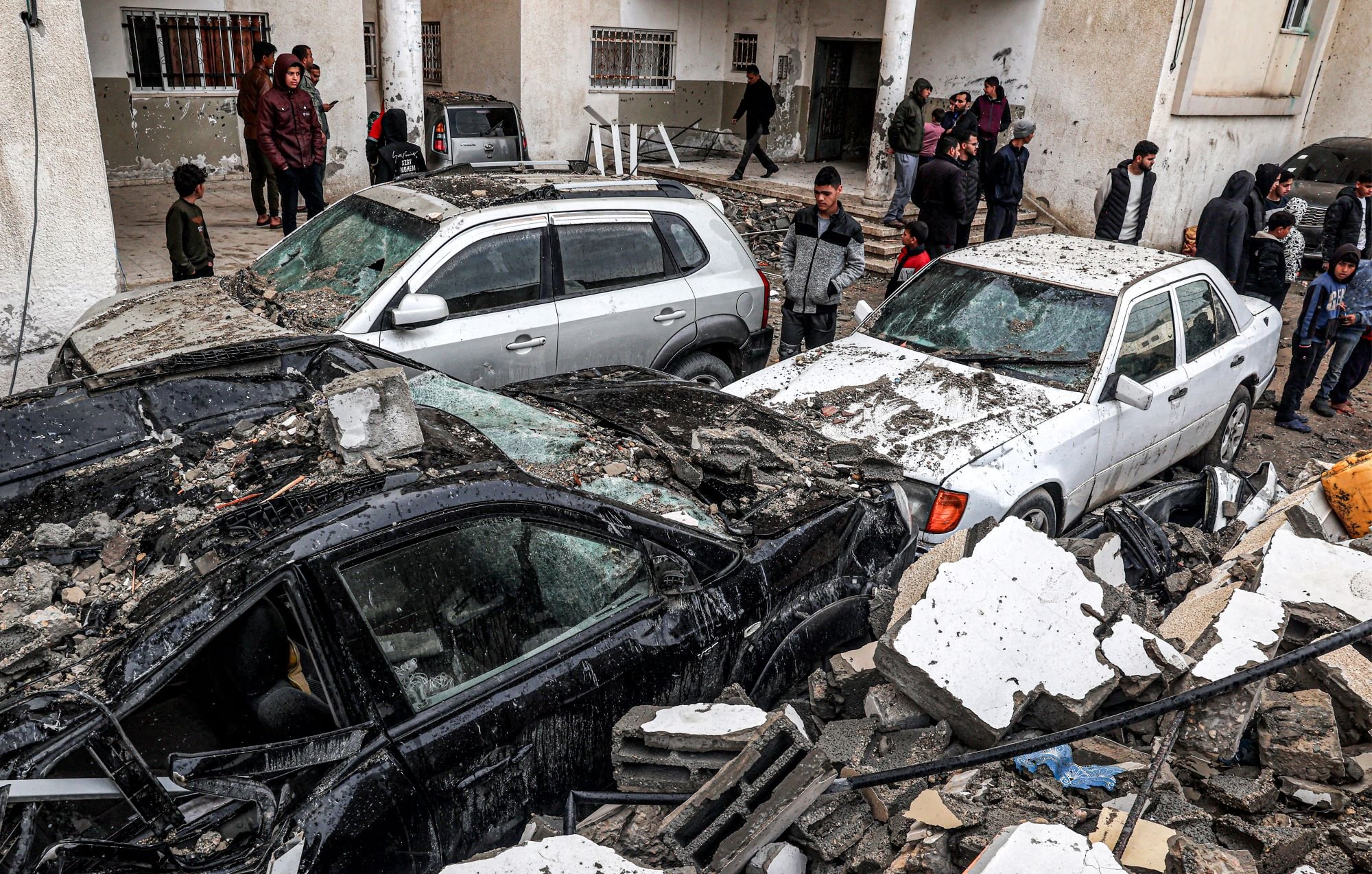 این تصویر نشاندهنده یک صحنه ویران شده به واسطه زلزله است. چندین خودرو زیر آوارهای ساختمانی قرار گرفتهاند و آسیب زیادی دیدهاند. افرادی در پسزمینه در حال مشاهده و بررسی وضعیت هستند. آوار خانهها و دیوارهای فروریخته در اطراف خودروها به چشم میخورد. به طور کلی، جو تصویر نشاندهنده اضطراب و فاجعه ناشی از زلزله است.