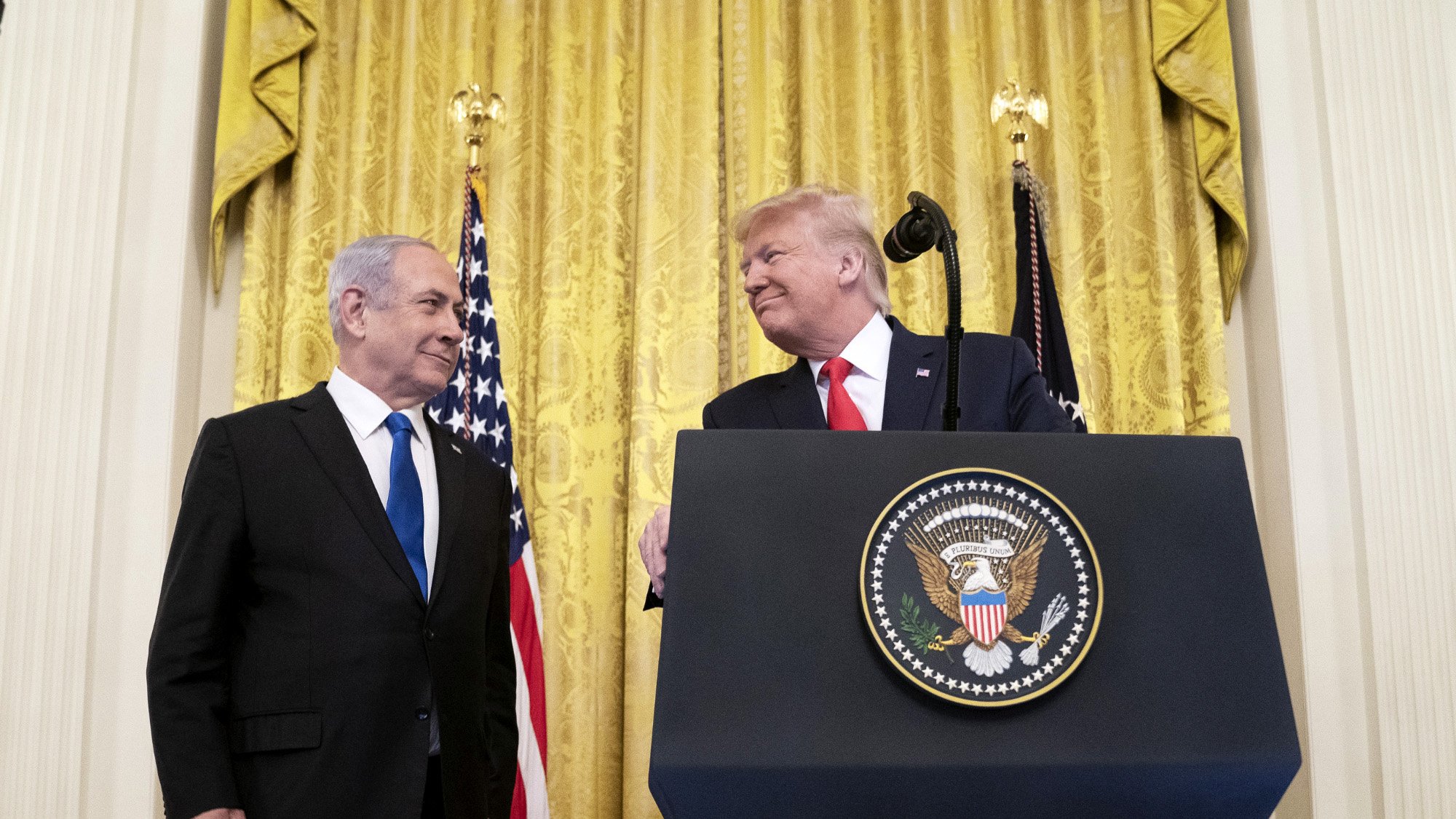 تصویر مربوط به یک مراسم رسمی است که در آن دو نفر در حال گفتگو هستند. در پسزمینه، پردههای طلایی و پرچمهای ایالات متحده دیده میشوند. یکی از افراد در سمت چپ، لباسی تیره به تن دارد و به شخص دیگر در سمت راست نگاه میکند. شخص در سمت راست یک لباس رسمی و یک کراوات به تن دارد و به نظر میرسد در حال لبخند زدن است. همچنین بر روی میزی که در مقابل آنها قرار دارد، نشان رسمی ریاستجمهوری ایالات متحده وجود دارد. این تصویر به نظر میرسد به یک رویداد سیاسی یا دیپلماتیک مربوط باشد.