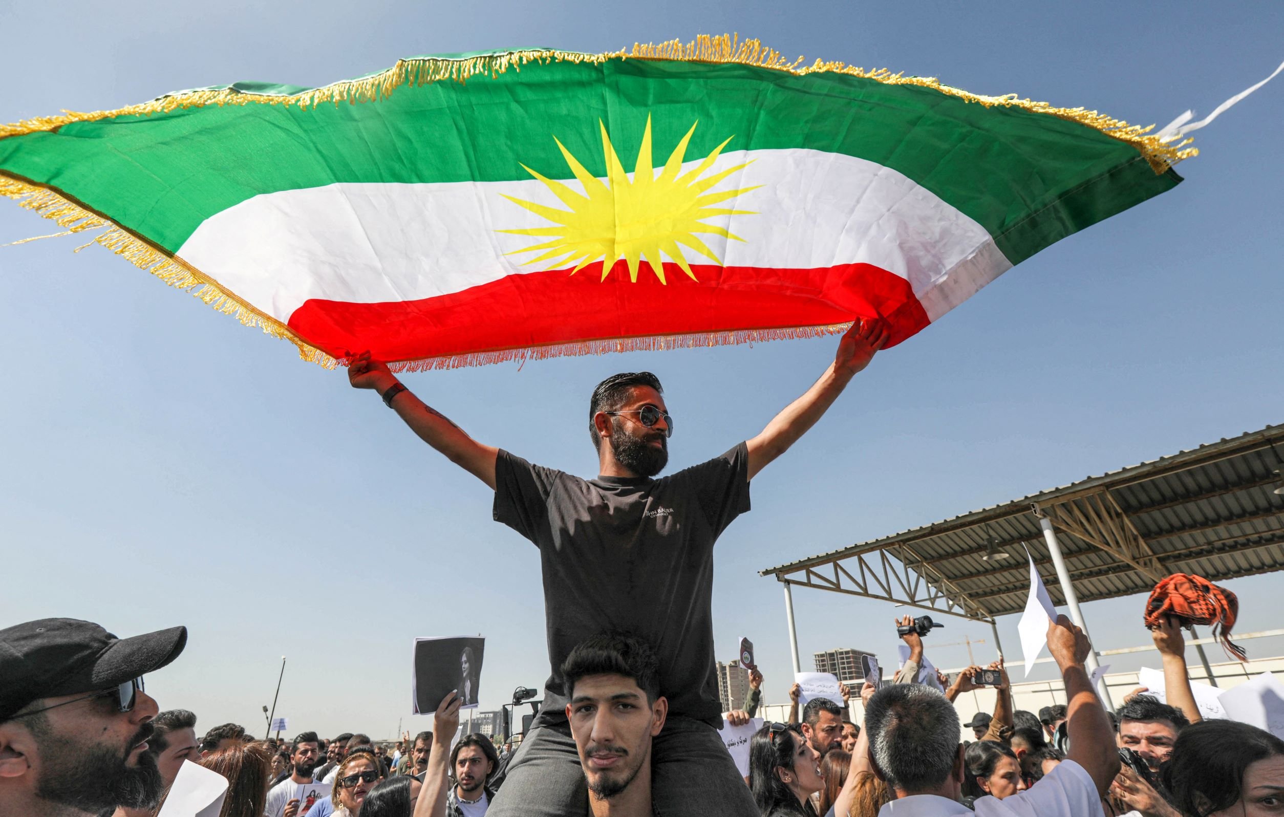 در این تصویر، فردی را مشاهده میکنیم که در بالای شانههای یک شخص دیگر ایستاده و پرچم کردستان را به اهتزاز درآورده است. پرچم دارای رنگهای سبز، سفید و قرمز با خورشید زرد در وسط است. جمعیتی در اطراف آنها حضور دارد و به نظر میرسد در حال برگزاری یک تظاهرات یا تجمع هستند. فضای تصویر آکنده از انرژی و احساسات جمعی است.