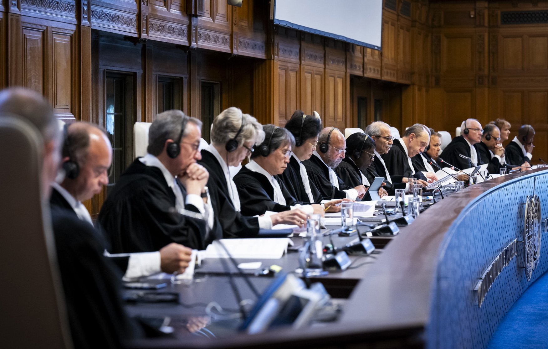 تصویر گروهی از قضات را نشان میدهد که در یک جلسه رسمی نشستهاند. آنها لباسهای مشکی قضائی به تن دارند و بسیاری از آنها هدفونهایی بر سر دارند. سالن دارای دکوراسیون چوبی و شکلی کلاسیک است و یک میز بزرگ به شکل نیمدایره در مرکز تصویر وجود دارد. قضات در حال بحث یا مطالعه اسناد هستند و فضای جدی و رسمی حاکم بر جلسه احساس میشود.