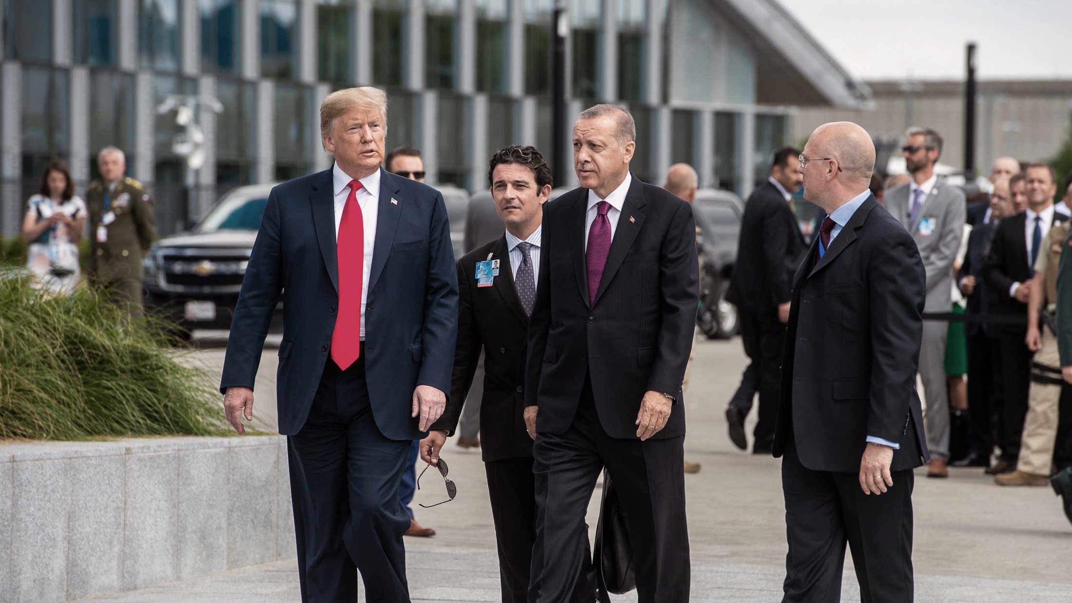 در این تصویر، چند مرد در حال پیادهروی هستند. یکی از آنها که لباس رسمی پوشیده و کلاهی تیره به سر دارد، به سمت دیگران نگاه میکند. شخص دیگری که در سمت چپ او قرار دارد، کت و شلوار و یک کراوات قرمز به تن دارد. دو مرد دیگر نیز به آنها ملحق شدهاند و ظاهراً در حال گفتگو هستند. پسزمینه، شامل ساختمانهای مدرن و تعدادی از افراد دیگر در حال حرکت است. جو کلی تصویر نشاندهنده یک ملاقات رسمی است.