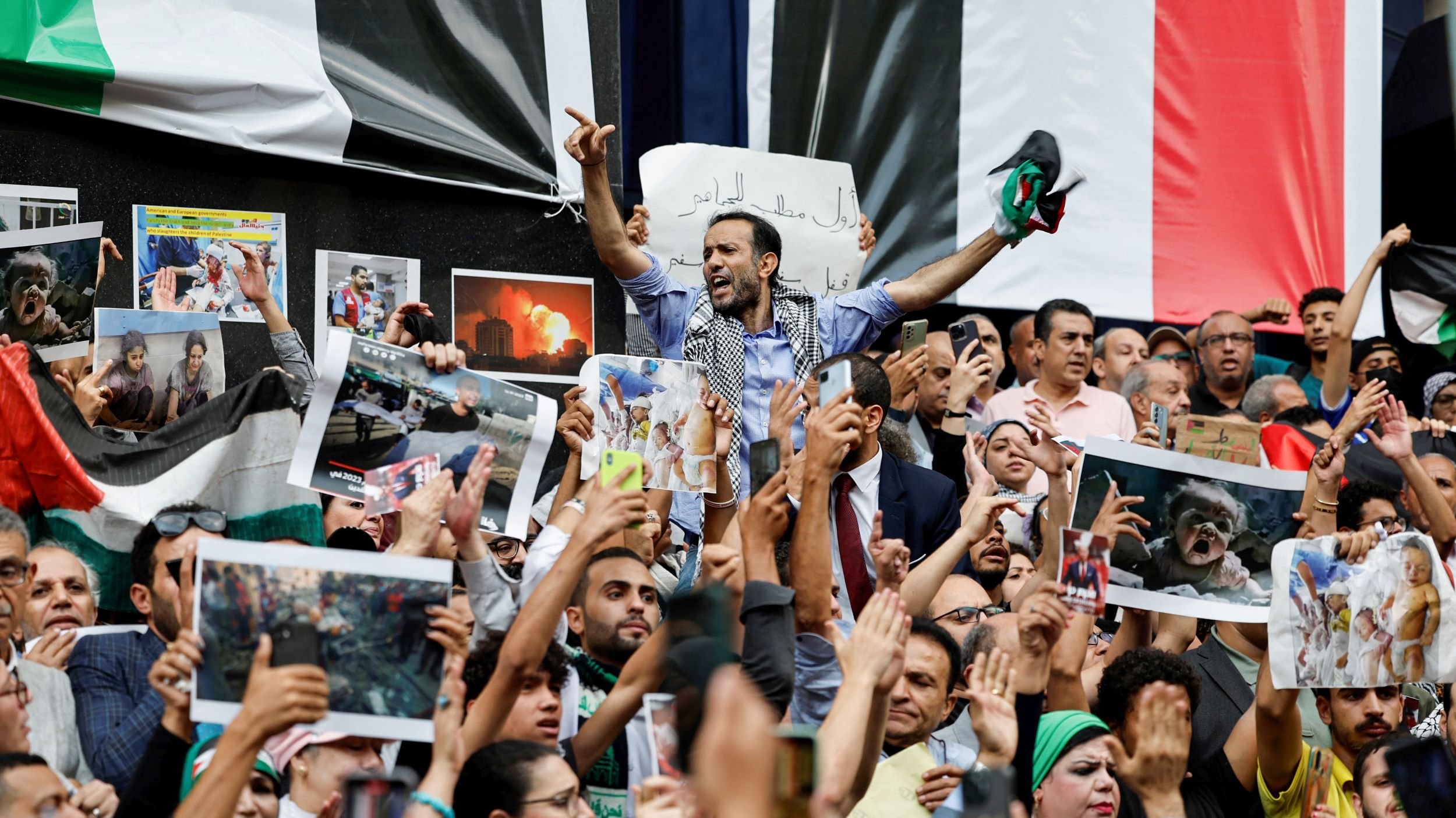 این تصویر یک تجمع بزرگ را نشان میدهد که مردم در آن با شور و هیجان حضور دارند. تعدادی از افراد در حال نشان دادن تصاویر و پلاکاردهایی هستند. برخی از معترضان در حال فریاد زدن یا ابراز احساسات خود هستند و مشخص است که موضوعی برای آنها اهمیت دارد. در پسزمینه، پرچمها و تصاویری از وقایع یا شخصیتهای مهم ممکن است مشاهده شود. جو کلی تصویر پر از انرژی و احساسات قوی است.