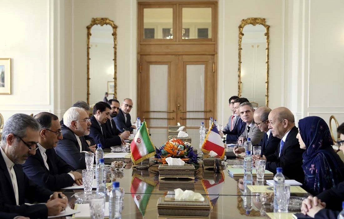 در این تصویر، یک جلسه رسمی در حال برگزاری است. تعدادی از افراد در دو طرف یک میز بزرگ نشستهاند و در مرکز میز، پرچمهای ایران و یک کشور دیگر قرار دارد. برخی از افراد به نظر میرسد که مقامات سیاسی یا دیپلمات هستند. فضا شبیه به یک سالن کنفرانس است، با نور طبیعی که از پنجرهها وارد میشود و دکوراسیون کلاسیک شامل آینهها و تابلوهای هنری. عدهای از افراد با دقت به سخنران گوش میدهند، در حالیکه برخی دیگر مشغول نوشتن یا یادداشتبرداری هستند.