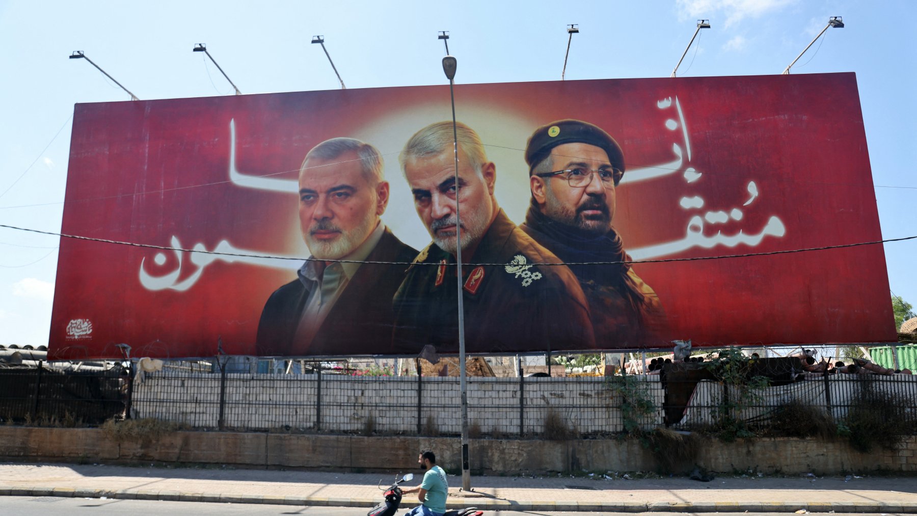 این تصویر یک بیلبورد بزرگ را نشان میدهد که سه نفر را به تصویر کشیده است. پسزمینه بیلبورد به رنگ قرمز است و ظاهراً به یک موضوع ملی یا سیاسی اشاره دارد. افراد تصویر شده به صورت نیمهتنه و با چهرههای جدی نمایش داده شدهاند. در پایین تصویر، یک موتور سوار در حال عبور است که نشاندهنده فعالیتهای روزمره در خیابان است. این بیلبورد ممکن است به یک رویداد خاص یا وحدت اشاره داشته باشد.