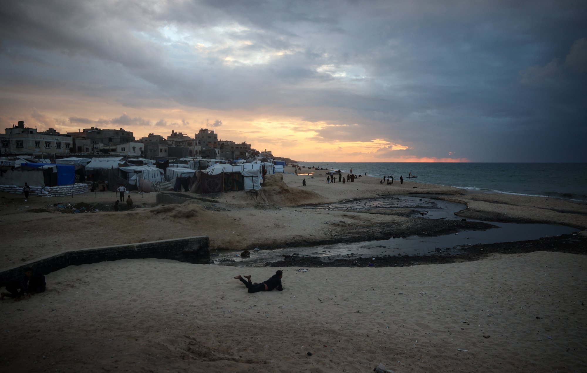تصویر نشاندهندهی یک ساحل است که در آن آسمان پهناوری با ابرهای تیره و روشن به تصویر کشیده شده است. خورشید در حال غروب است و نور ملایمی بر روی آب دریا و شنهای ساحل میتابد. در سمت چپ تصویر، برخی چادرها و سازههایی دیده میشود که نشاندهندهی سکونتگاههای موقت هستند. همچنین، در دوردست، افرادی در حال حرکت و فعالیت هستند. این تصویر حال و هوای خاصی از مکانهای ساحلی همراه با چالشهای اجتماعی و محیطی را منتقل میکند.