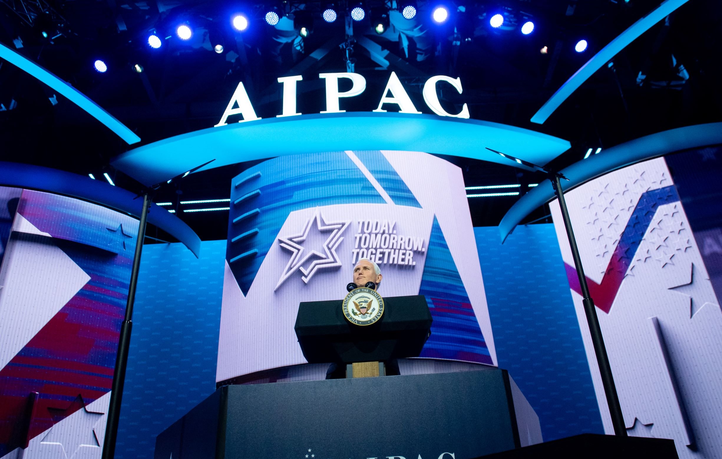 تصویر یک صحنه از یک کنفرانس یا گردهمایی است. در پسزمینه، یک نمایشگر بزرگ با نام "AIPAC" و شعار "Together Tomorrow" دیده میشود. در جلوی صحنه، یک میز سخنرانی وجود دارد که بر روی آن نشان رسمی (احتمالاً نشان کشور یا دولت) قرار دارد. نورپردازی آبی و سفید به فضایی بانشاط و رسمی بخشیده است. طراحی کلی صحنه نشان دهنده یک رویداد مهم است.