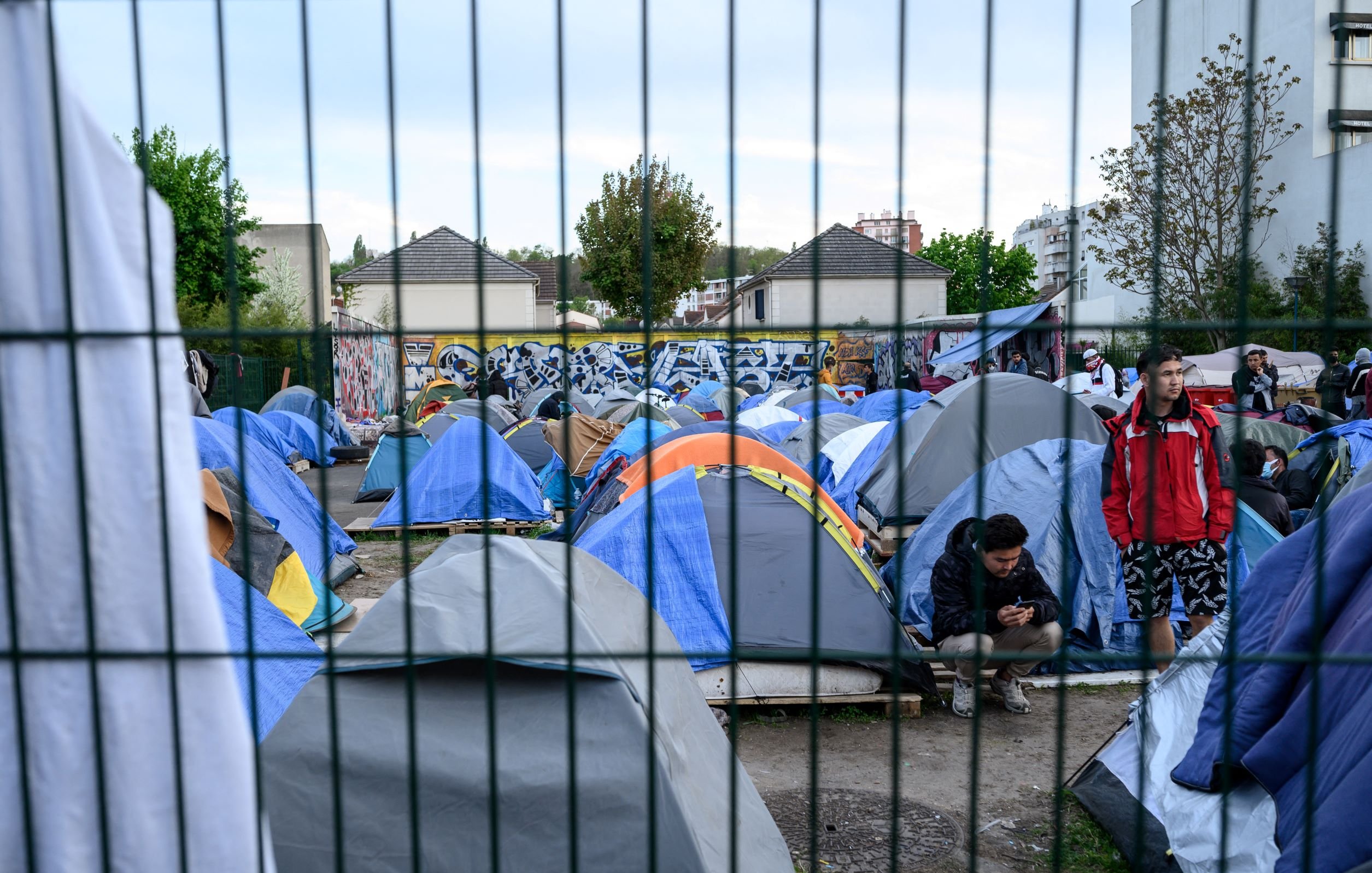 این تصویر نمایی از یک اردوگاه چادرها را نشان میدهد. چادرهای متعدد با رنگهای مختلف در یک فضای باز قرار دارند و برخی از افراد در اطراف آنها مشغول فعالیت هستند. در پسزمینه، یک دیوار با هنر خیابانی یا گرافیتی قابل مشاهده است. همچنین، یک نرده فلزی در جلوی تصویر وجود دارد که فضا را از محیط بیرونی جدا میکند. هوای آسمان میتواند کمی ابری به نظر برسد.
