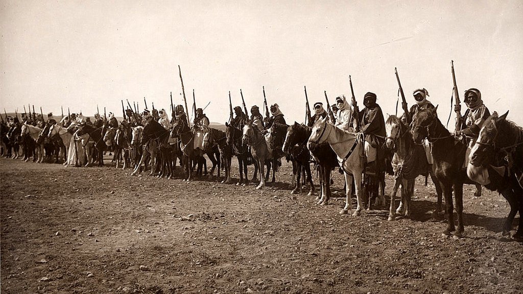 این تصویر نشاندهنده گروهی از سواران را در یک خط منظم است. آنها بر روی اسبهای خود نشستهاند و برخی از آنها نیز نیزه یا سلاحهایی در دست دارند. لباسهای آنها سنتی به نظر میرسد و فضایی تاریخی را القا میکند. زمین زیر پای آنها خشک و سنگی است و در پسزمینه، فضای باز و آسمان دیده میشود. این تصویر میتواند اشاره به یک دوره خاص از تاریخ یا مراسمی فرهنگی داشته باشد.